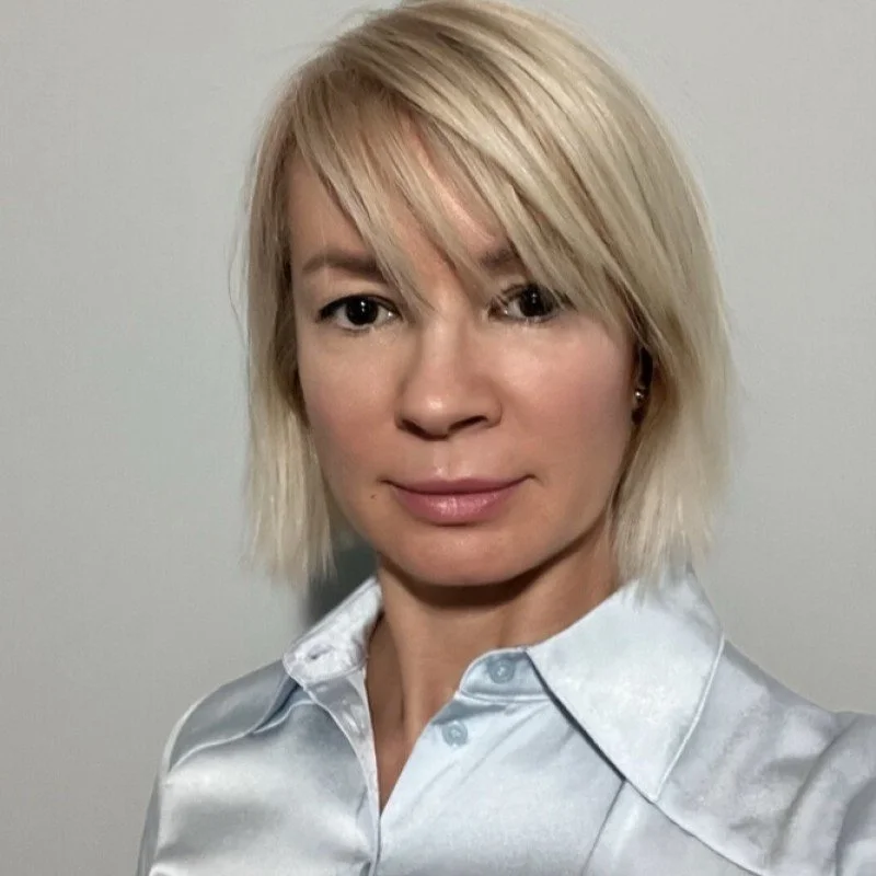 A woman with short blonde hair, wearing a white satin shirt, standing against a plain background.
