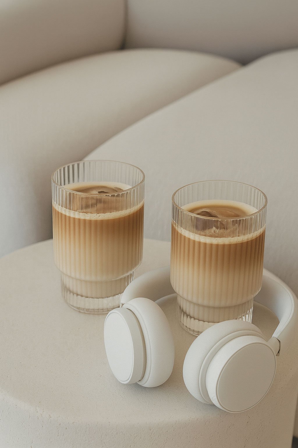 Two glasses of iced coffee and white headphones on a white surface against a light-colored upholstered background.