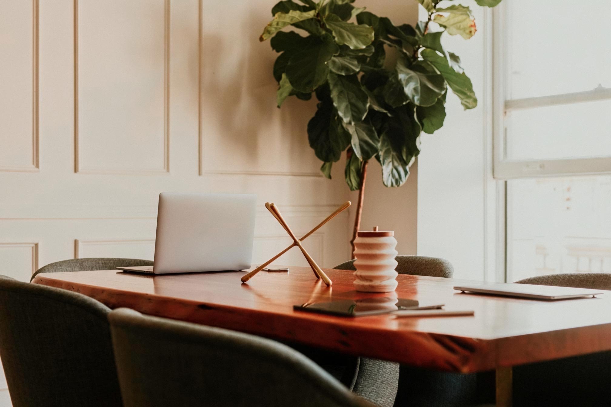 A wooden dining table with a laptop, a ceramic vase, and a notepad, surrounded by upholstered chairs. A large potted plant is in the background near a paneled wall and window.