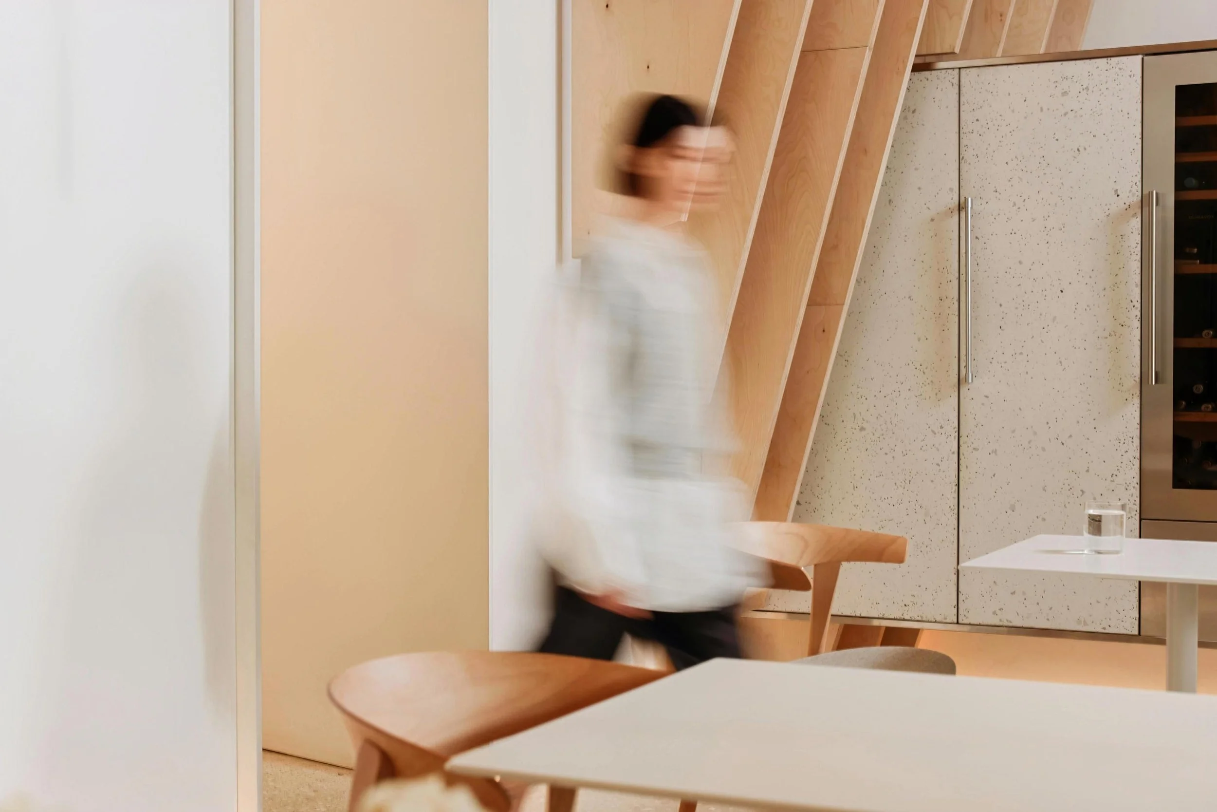 A blurred person walking past a modern interior with wooden shelves, a cabinet, and a white table in an art gallery or cafe.