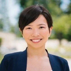 Portrait of an Asian woman with short black hair, smiling, wearing a navy blazer outdoors with blurred trees in the background.