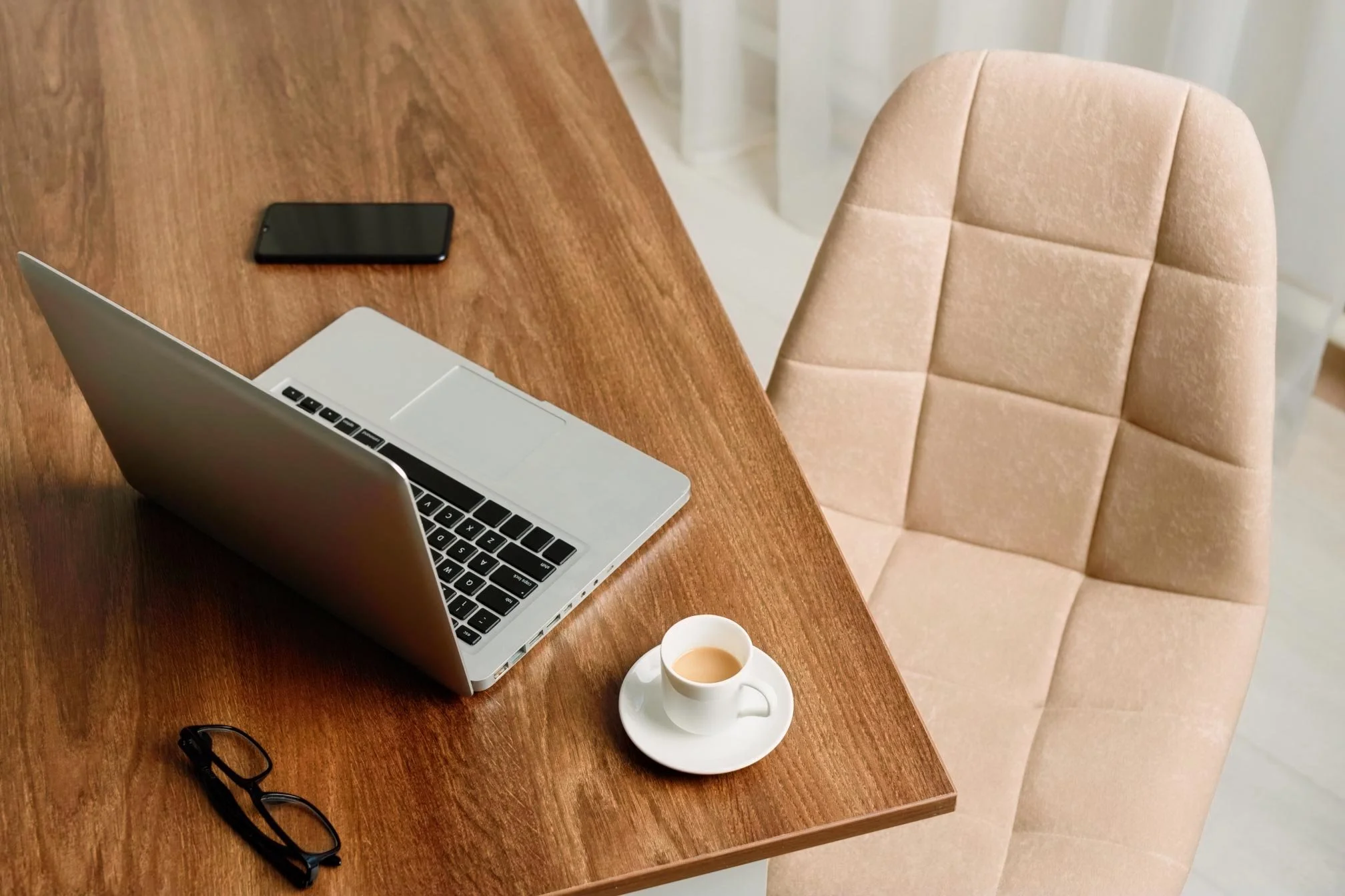 An open silver laptop, a smartphone, a pair of glasses, and a cup of coffee on a wooden table next to a beige upholstered chair.
