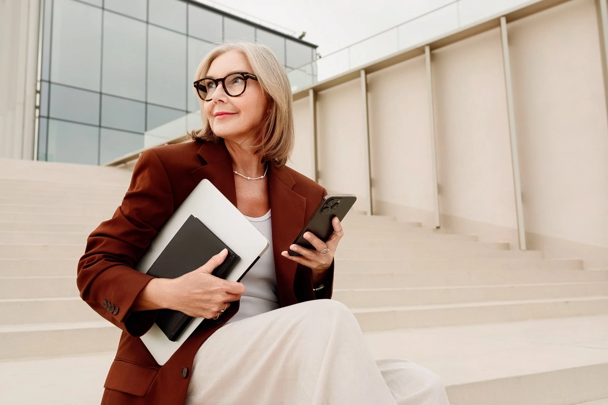 A middle-aged woman with gray hair, wearing glasses, a brown blazer, and beige pants, sitting on outdoor stairs near a modern glass building, holding a smartphone and a laptop.