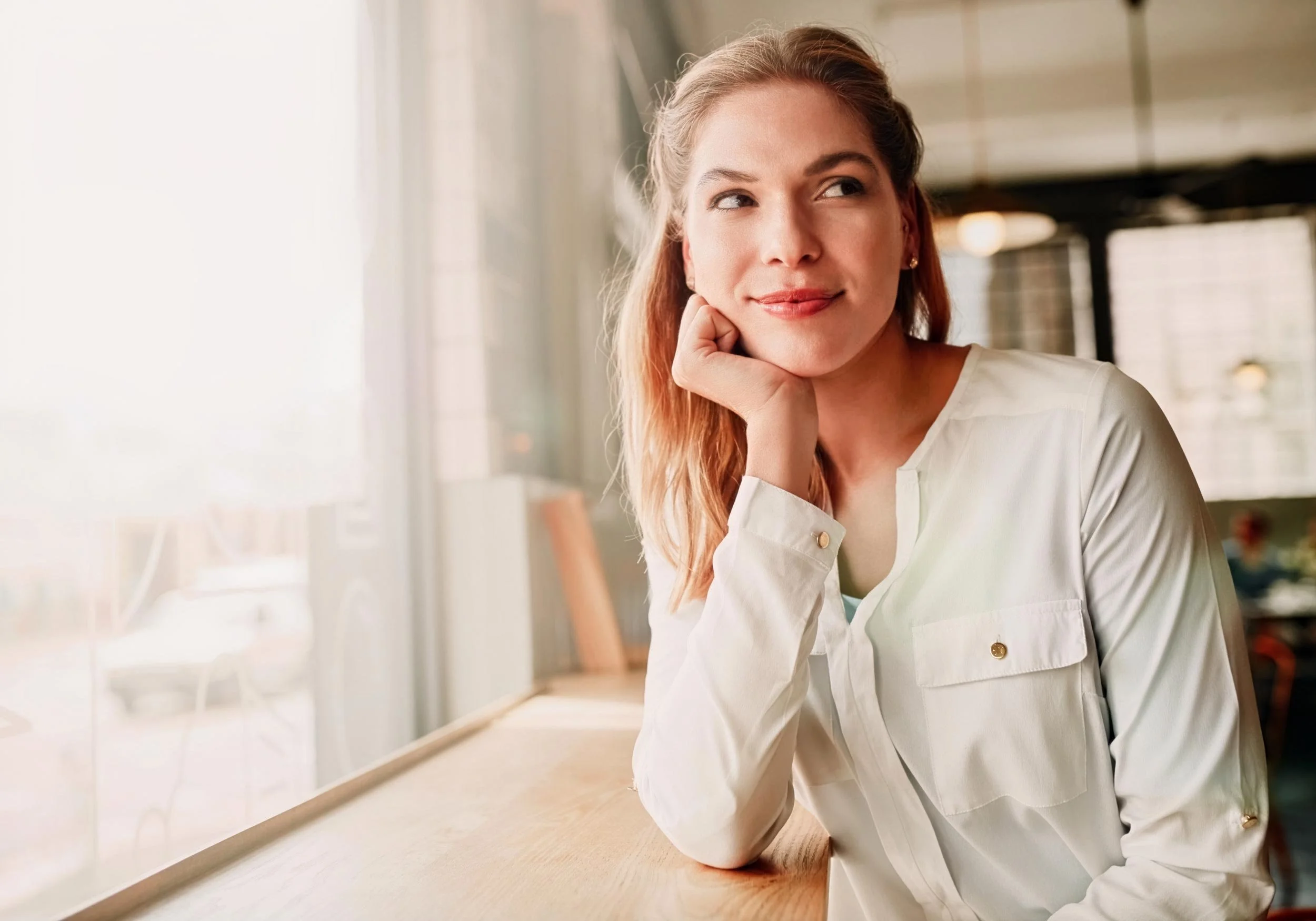 A woman with light brown hair, wearing a white shirt, sitting at a table in a bright, modern space with large windows and warm lighting, resting her chin on her hand and looking to her right with a slight smile.