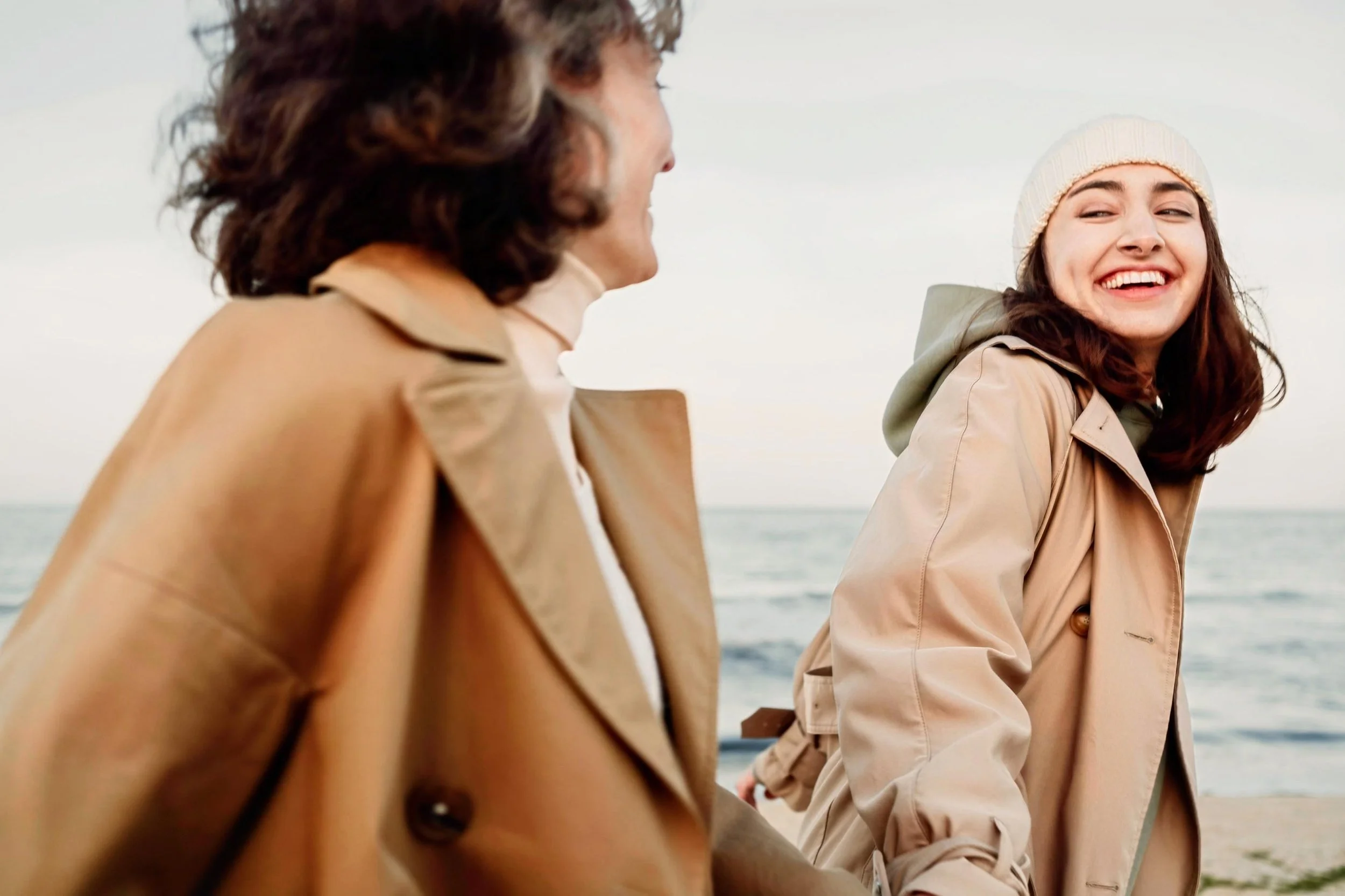 A young woman with brown hair wearing a beige coat and a white knit beanie smiling at a man with brown hair wearing a beige trench coat by the ocean.