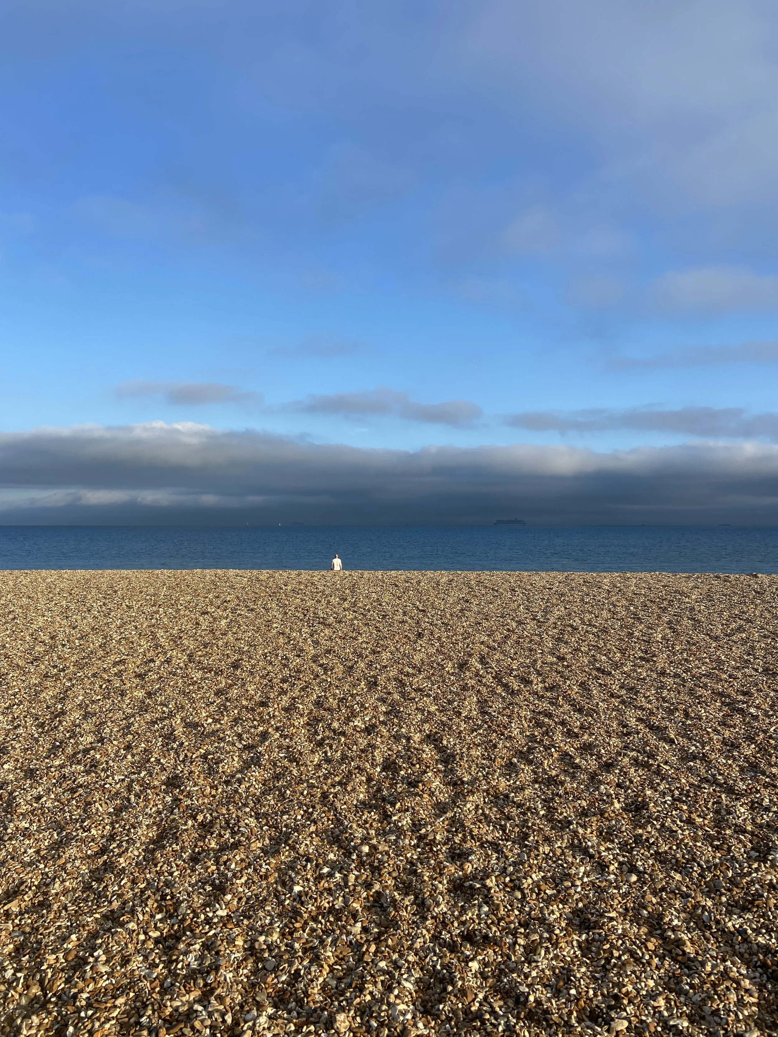 A person sitting on a pebble beach facing the ocean with blue sky and clouds overhead.