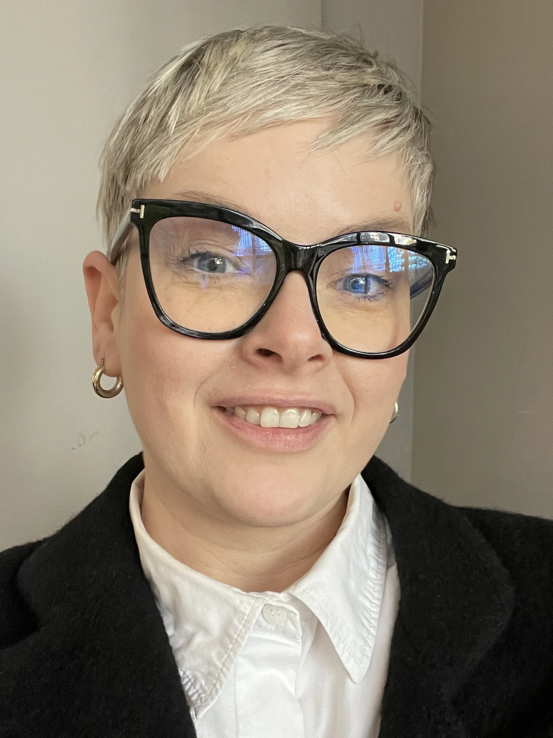 A woman with short blonde hair, wearing large black glasses, gold hoop earrings, and a white collared shirt under a black sweater, smiling at the camera.