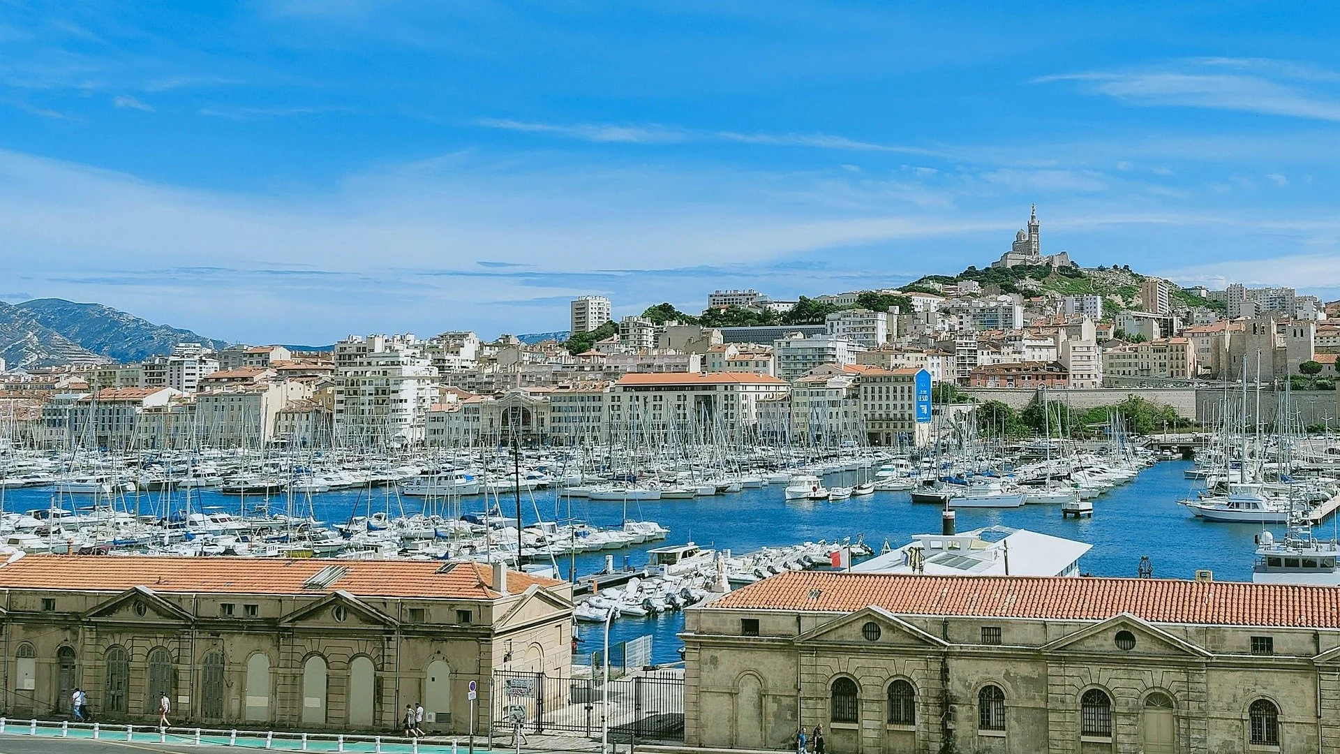 Vue du port de Marseille avec des yachts amarrés, bâtiments anciens et la basilique Notre-Dame de la Garde sur la colline en arrière-plan sous un ciel bleu.