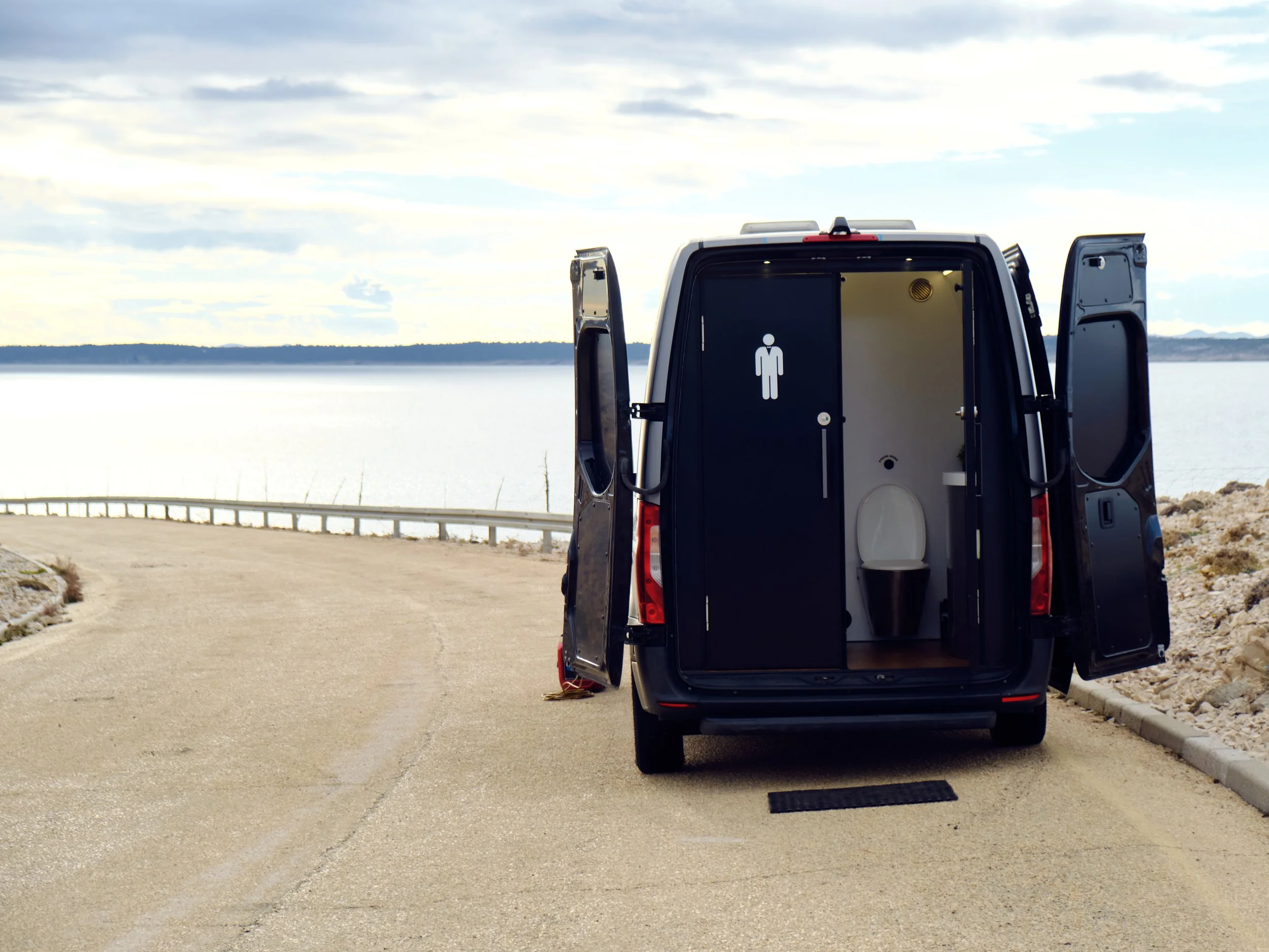 A van converted into a portable restroom with a man symbol, parked on a scenic road beside a body of water under a partly cloudy sky.