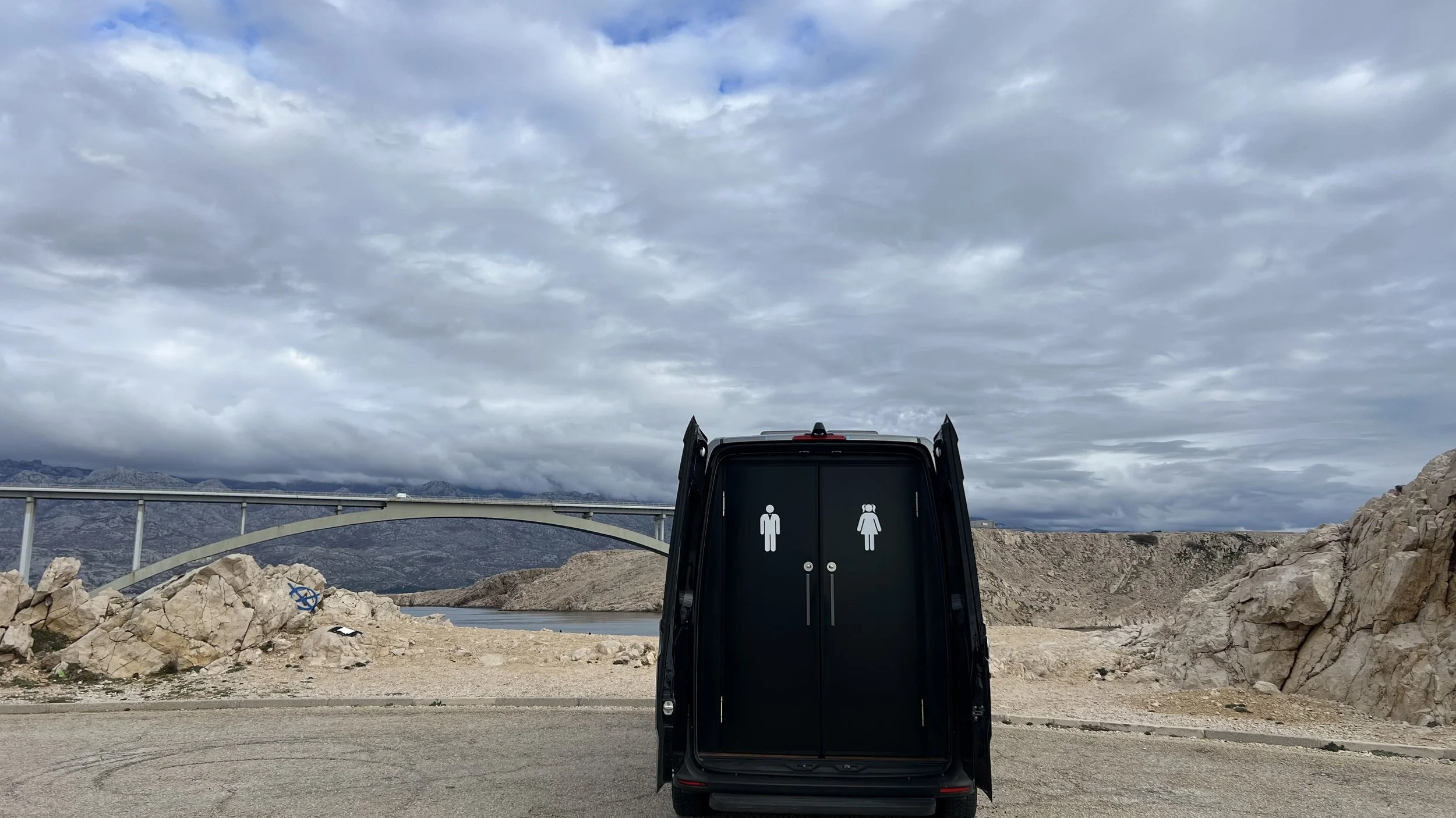 Public restroom with male and female symbols on black doors, set in a barren rocky landscape with mountains and a cloudy sky in the background.