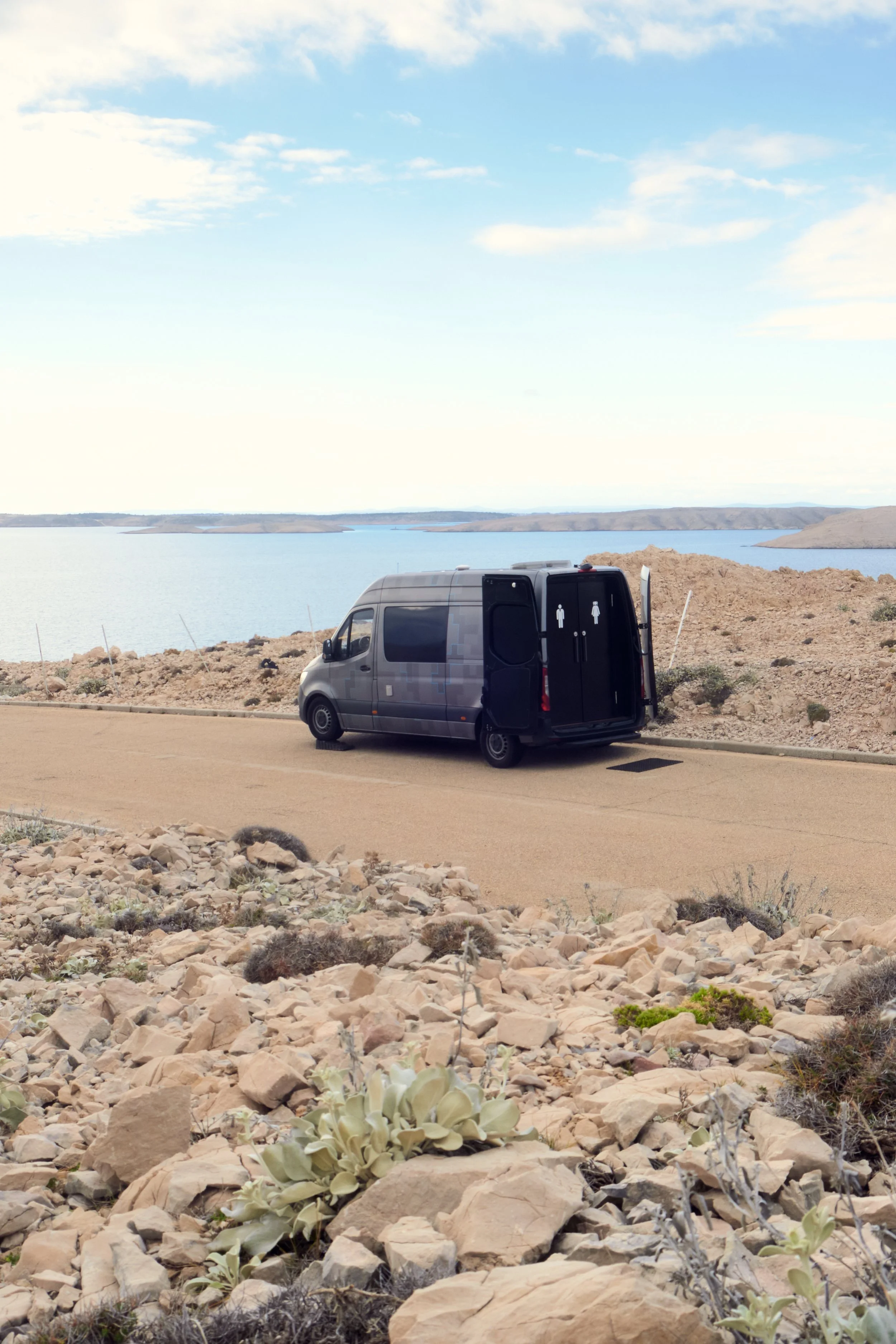 A black van parked on a roadside near a body of water with rocky terrain in the foreground and a partly cloudy blue sky above.