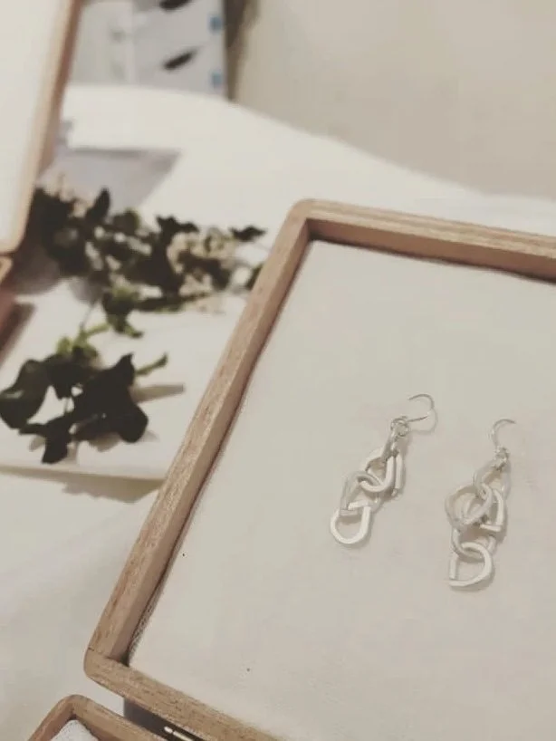 Boucles d'oreilles en métal argenté dans une boîte en bois, avec des fleurs séchées floues en arrière-plan.