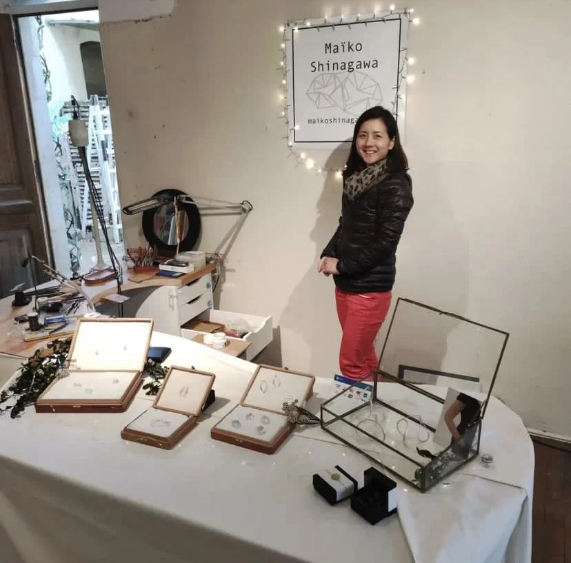 Femme souriante derrière une table avec des boîtes à bijoux et des bijoux exposés, dans un intérieur décoré avec une lumière de Noël et un panneau avec le nom Maïko Shinagawa.
