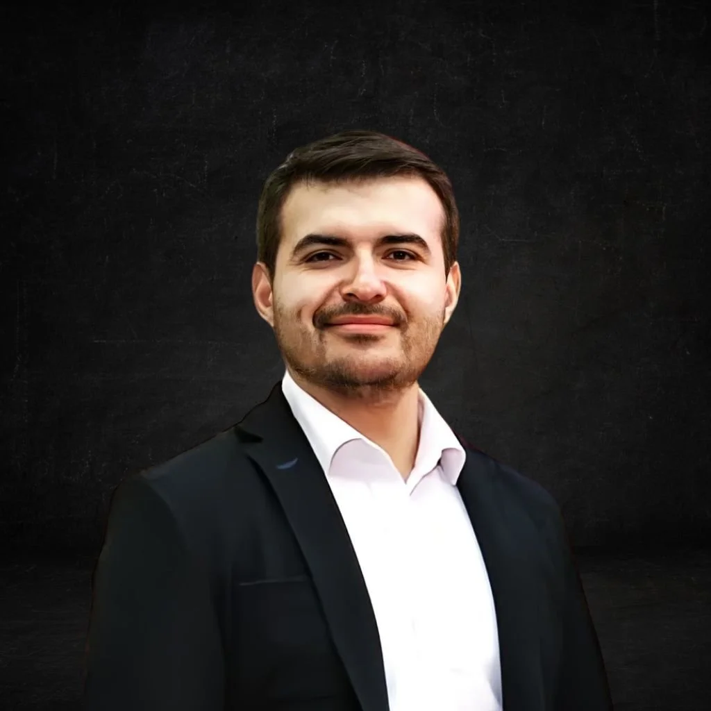 Retrato de un hombre con cabello castaño, barba bien recortada, vistiendo saco negro y camisa blanca, con fondo negro.