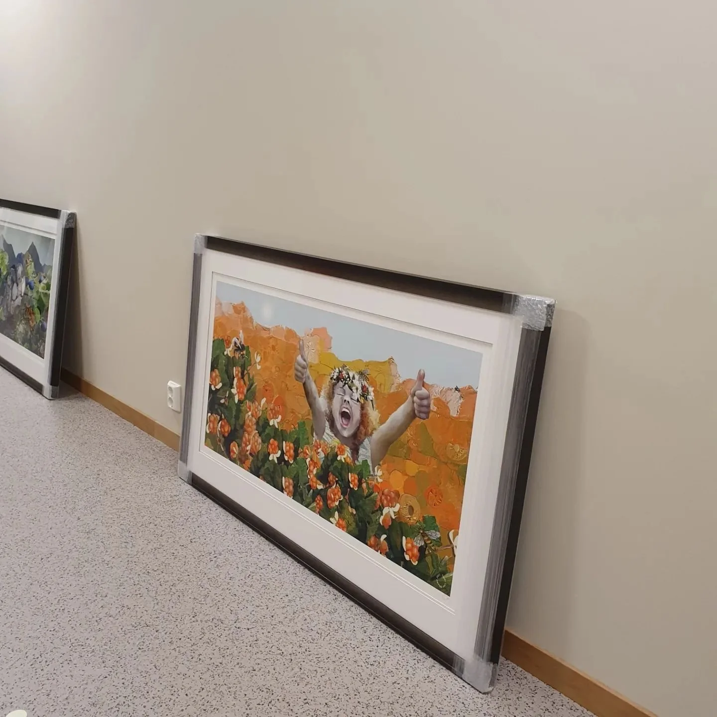 To bilderammer som er plassert mot en beige vegg, på gulvet, med innrammete kunstverk. Den ene rammen har et kunstverk av en person med blond hår og briller, som smiler og gir tommel opp mot en bakgrunn av orange fjell og grønne planter.