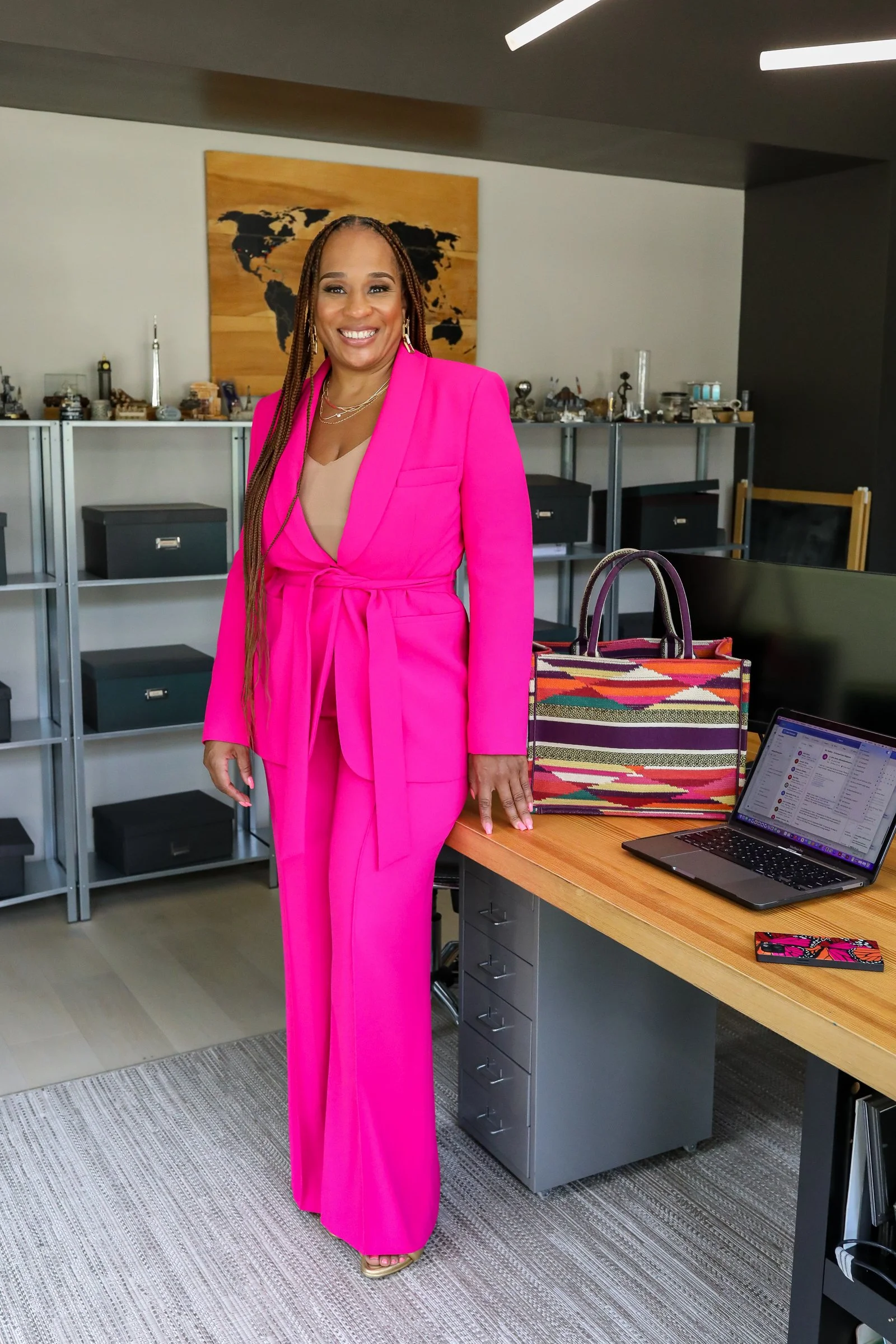 Founder, Ashley Page,  smiling with long braided hair wearing a bright pink suit with matching pants, standing in an office environment. Behind her is a world map artwork, and on the desk are colorful bags and a laptop.