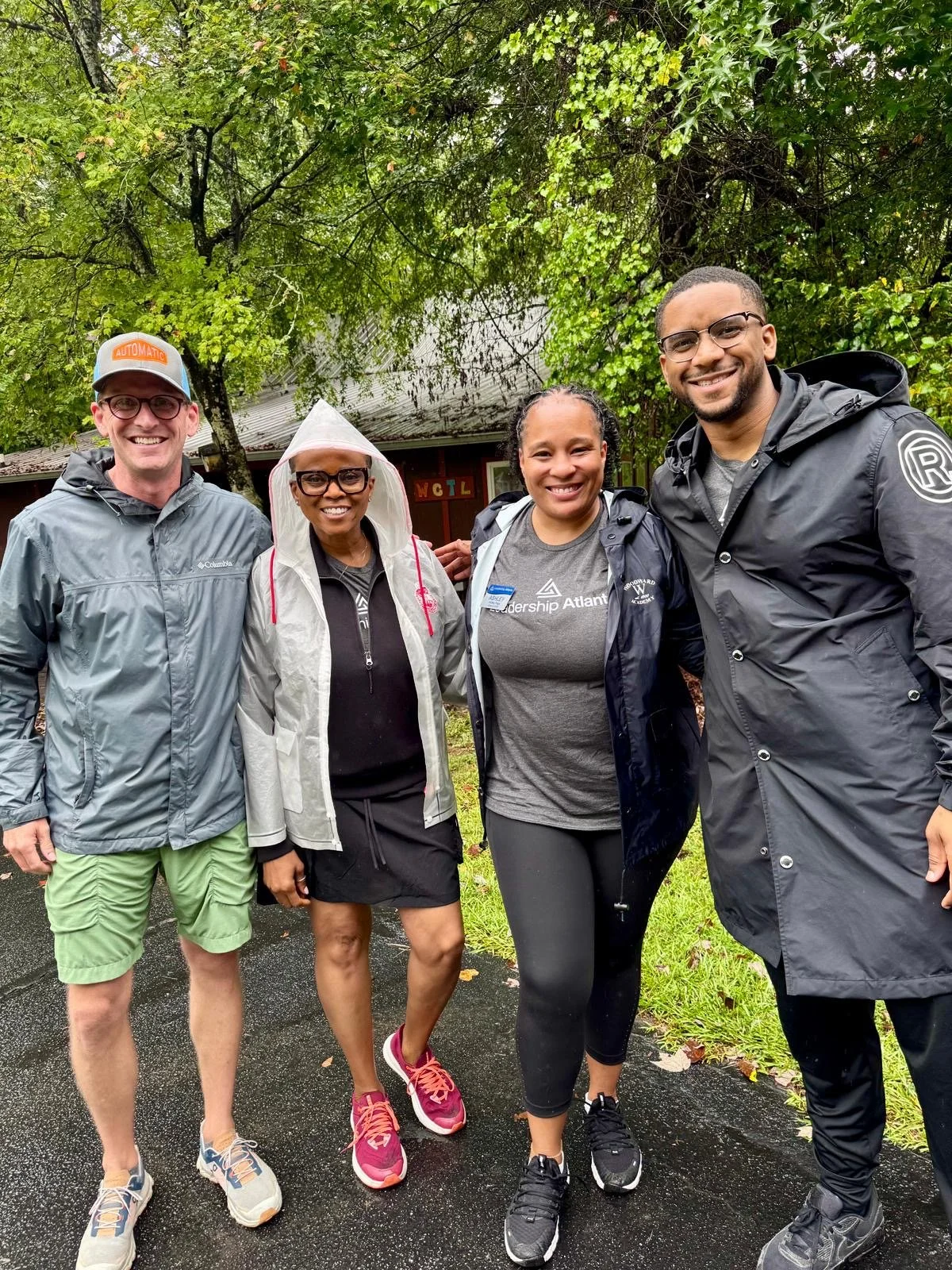 Ashley Page standing with Atlanta Leadership team outdoors on a rainy day, smiling, dressed in rain jackets and athletic clothing, with green trees and a building in the background.