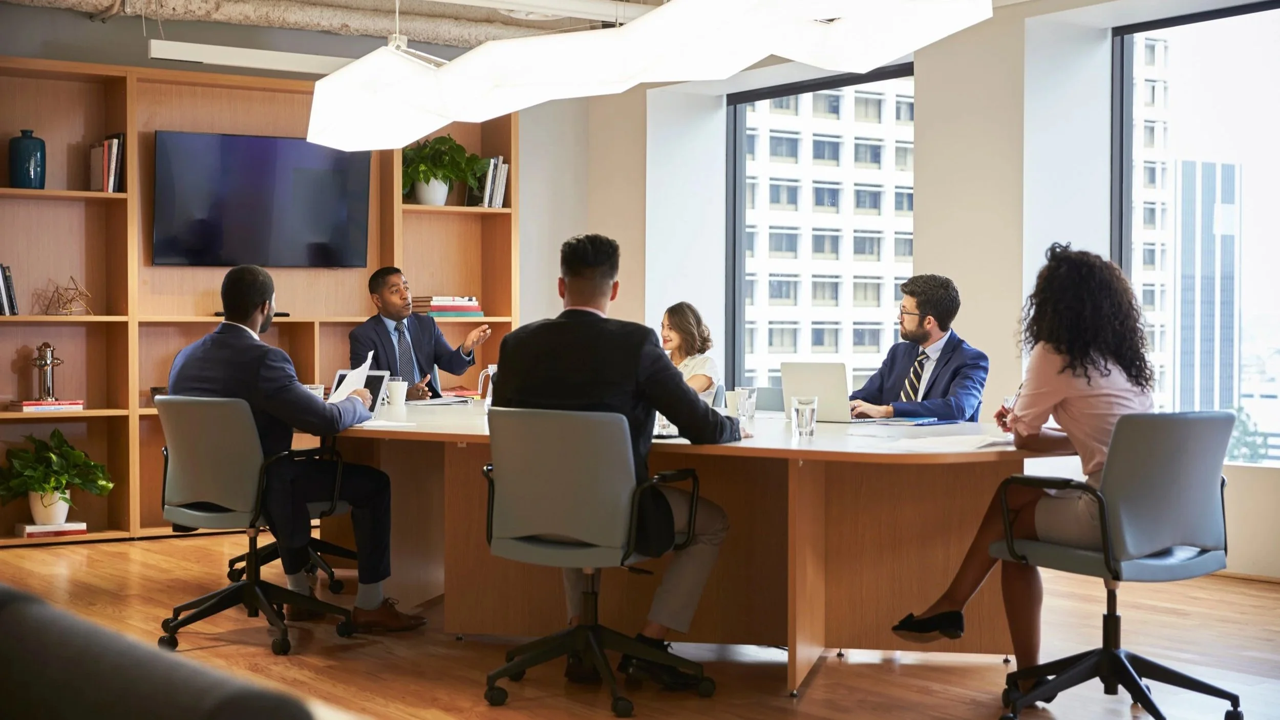 A business meeting with six diverse professionals seated around a large table in a modern office with large windows, bookshelves, and a large flat-screen TV on the wall. They appear to be engaged in discussion.