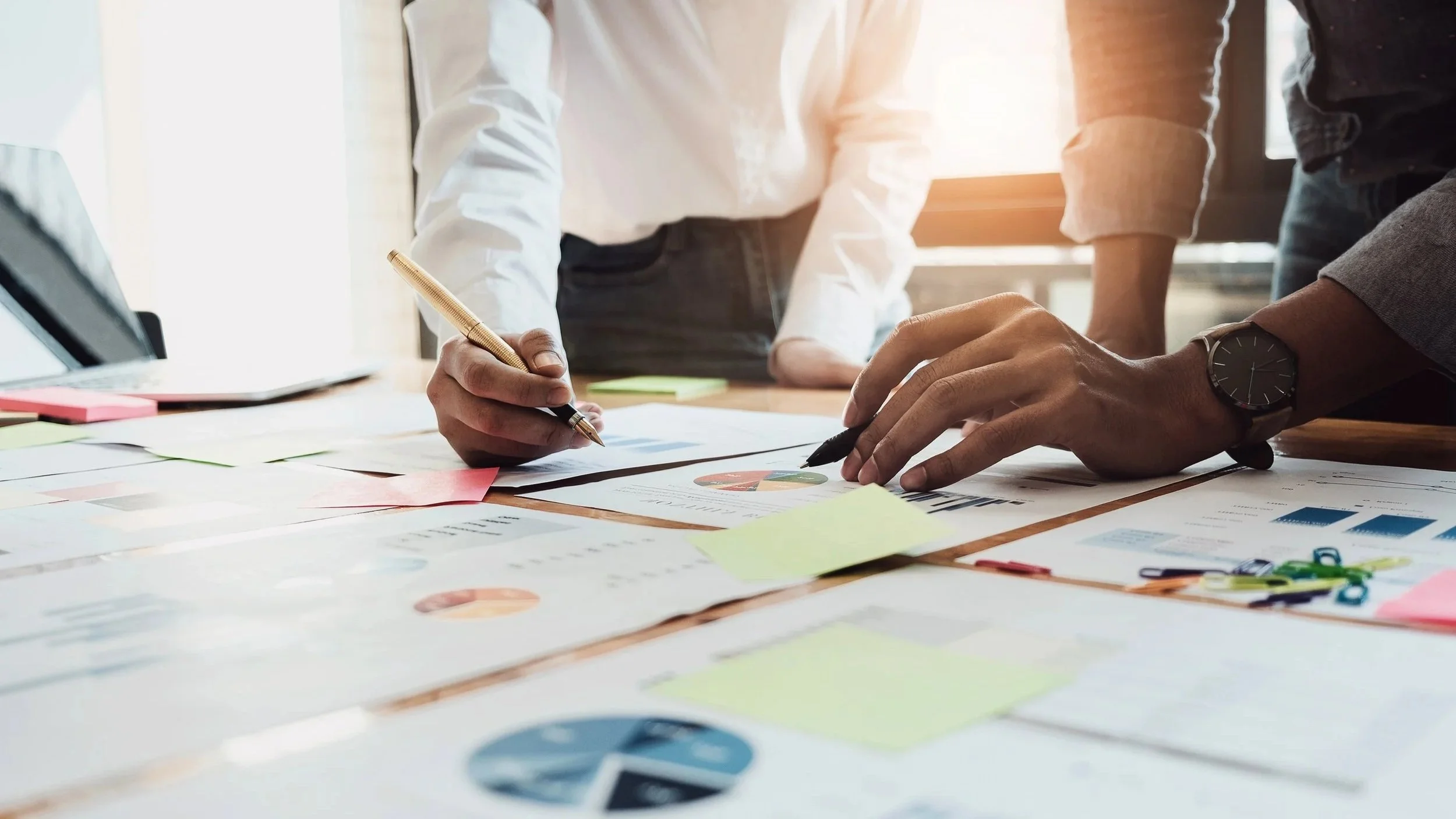 Two people are working at a cluttered table with documents and colorful sticky notes, using pens to review charts and graphs.