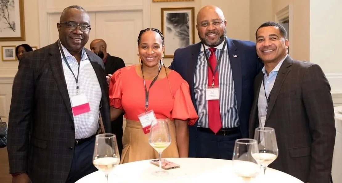 Ashley Page smiling and posing with 3 other people for a Legal professional social event, standing behind a table with three glasses of white wine.