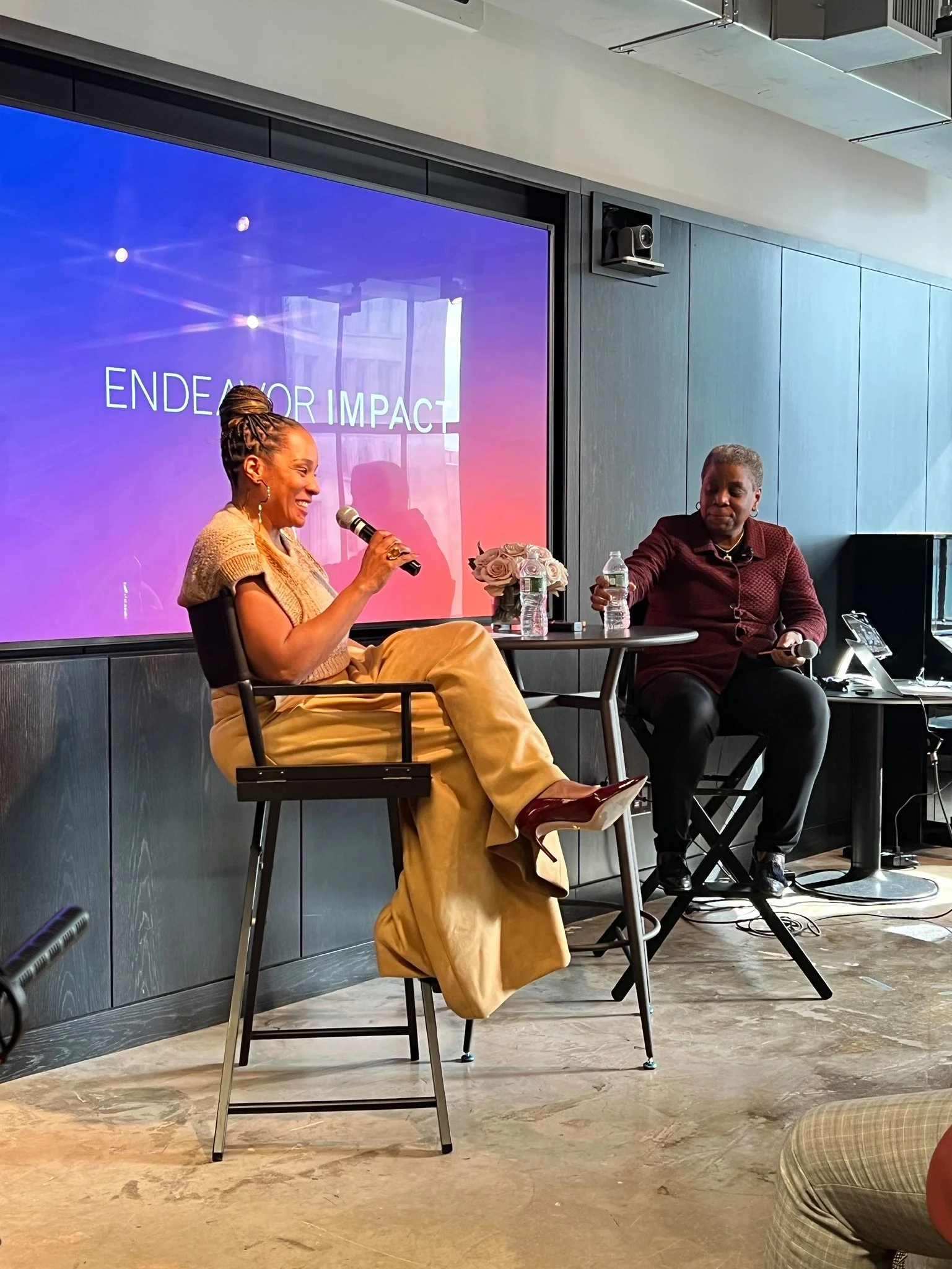 Two women sitting at a table during a discussion or interview, with a large screen behind them displaying the words "Endeavor Impact". One woman is speaking into a microphone, while the other listens attentively. There are water bottles, a small bouquet of roses, and some electronic equipment on the table.