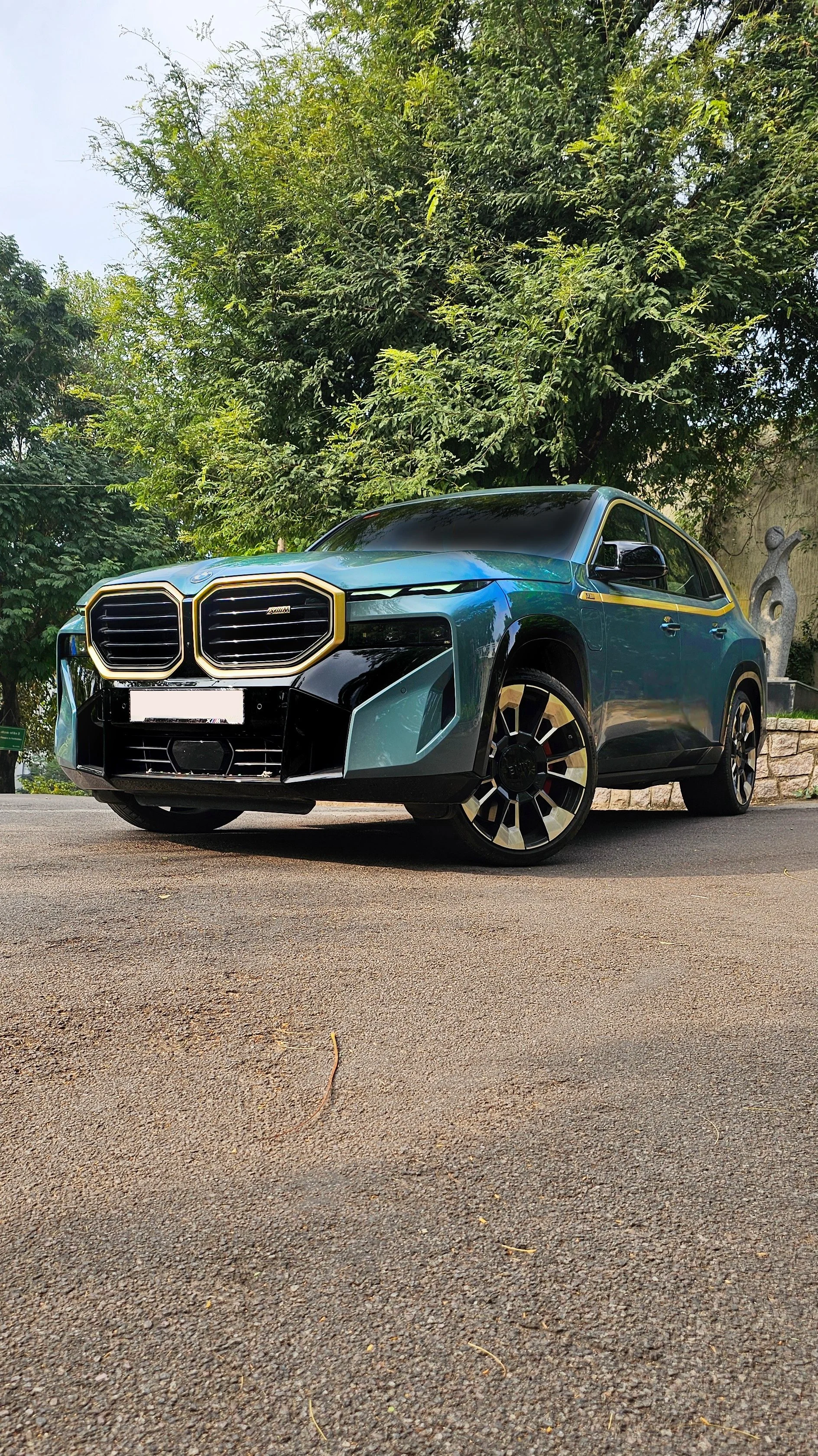 A blue BMW XM SUV with black and gold accents parked on a paved road, with trees and greenery in the background.