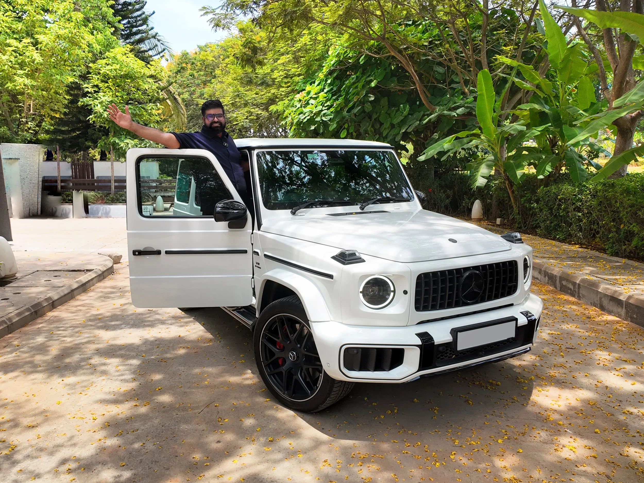 An Indian man with glasses, a beard, and wearing a dark shirt standing in the open door of a white Mercedes-Benz G Wagon SUV, waving, with lush green trees in the background.