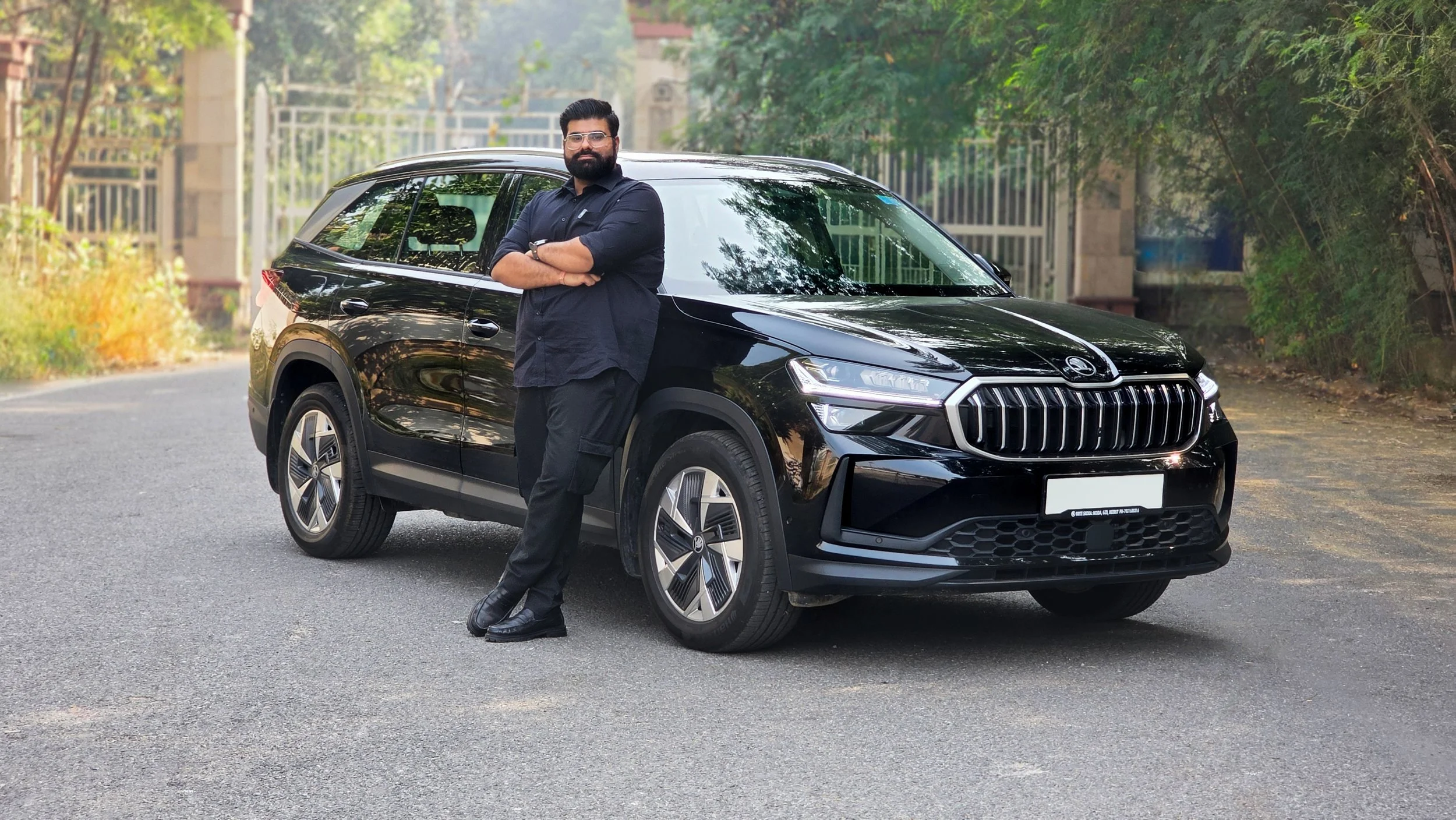 An Indian man with a beard and glasses wearing black shirt and pants, standing with arms crossed in front of a black Skoda Kodiaq SUV car on a paved road surrounded by trees.