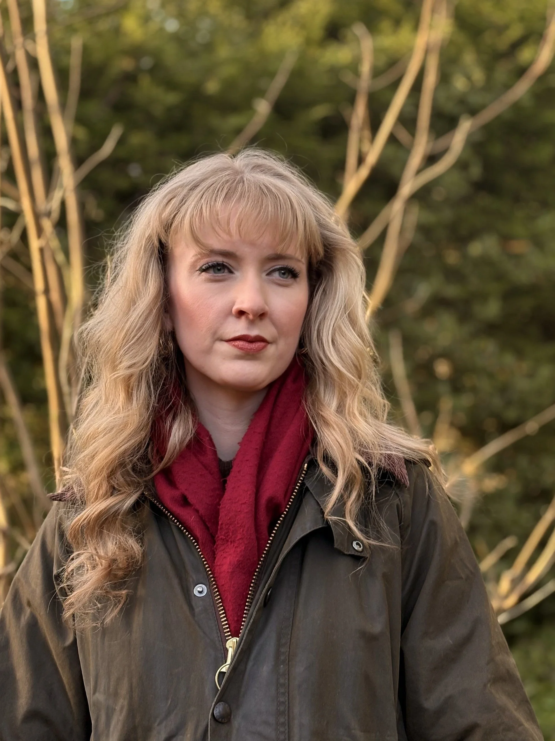 Madeleine Hall. She has wavy blonde hair and blue eyes, wearing a red scarf and a dark green jacket, standing outdoors with trees and branches in the background.