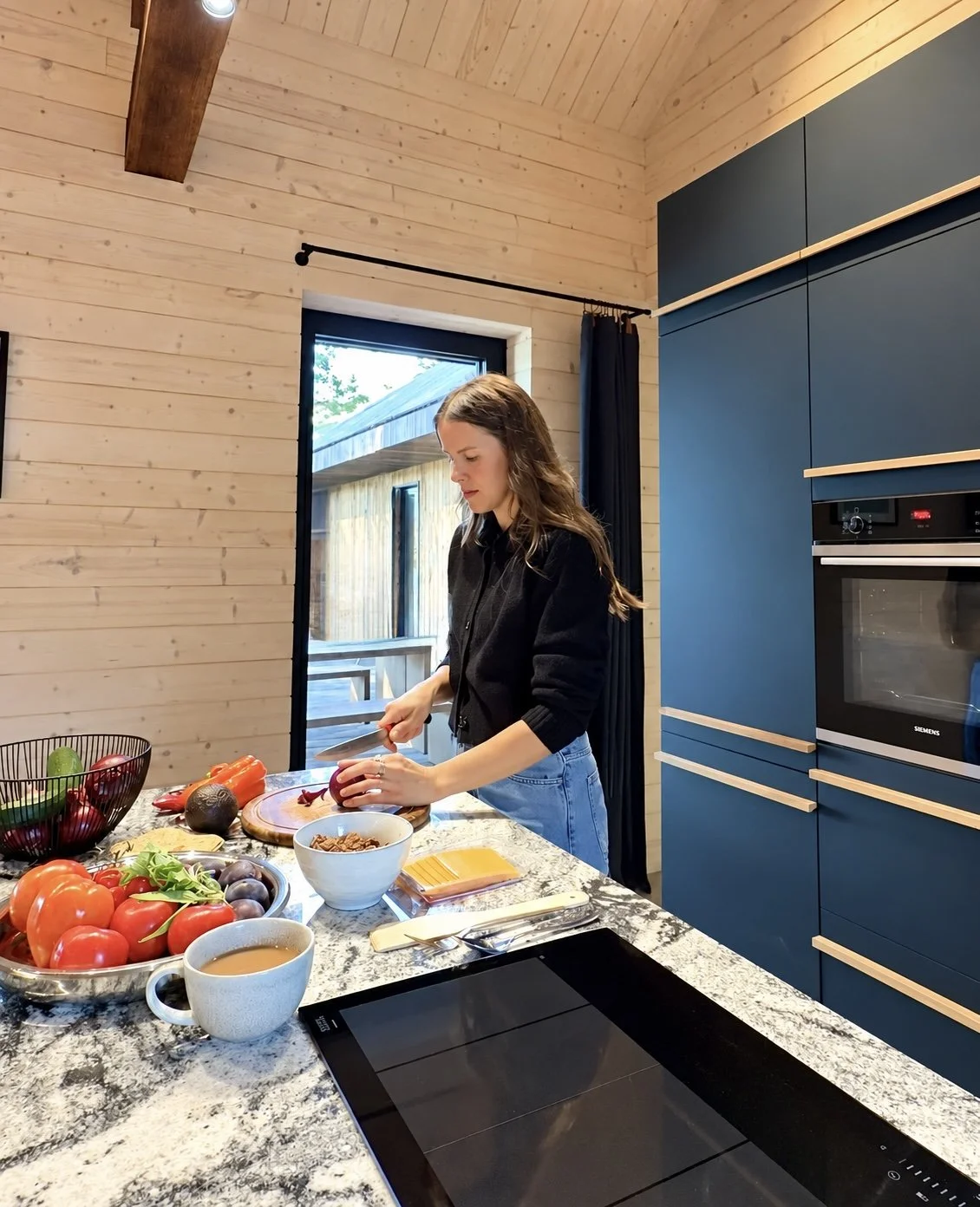 A woman with brown hair in a black shirt and blue jeans preparing vegetables in a modern kitchen with a wooden interior, blue cabinets, and a marble countertop, during daytime.