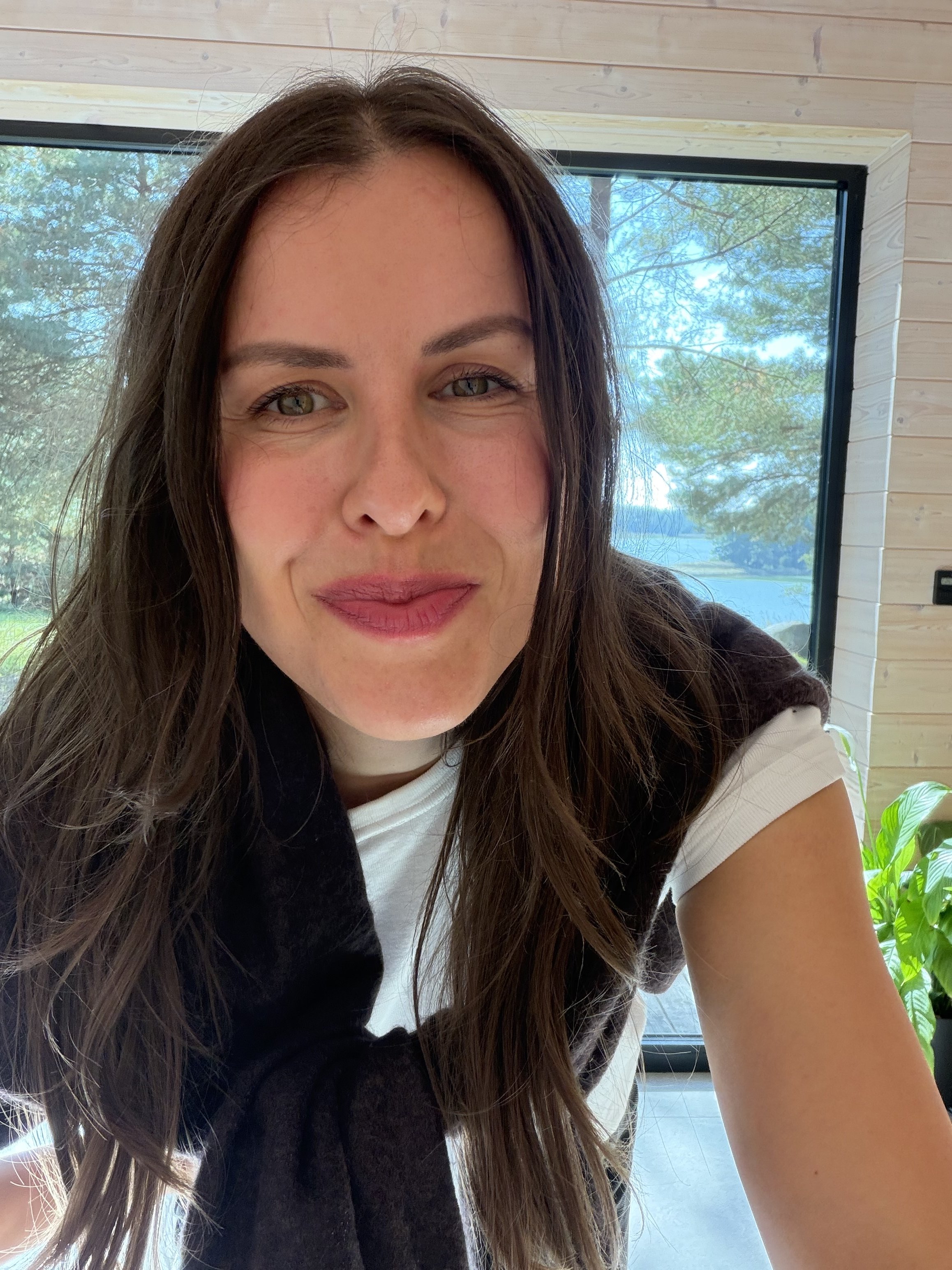 Close-up portrait of Anna Retel smiling, with long dark hair, in front of a window with trees behind her