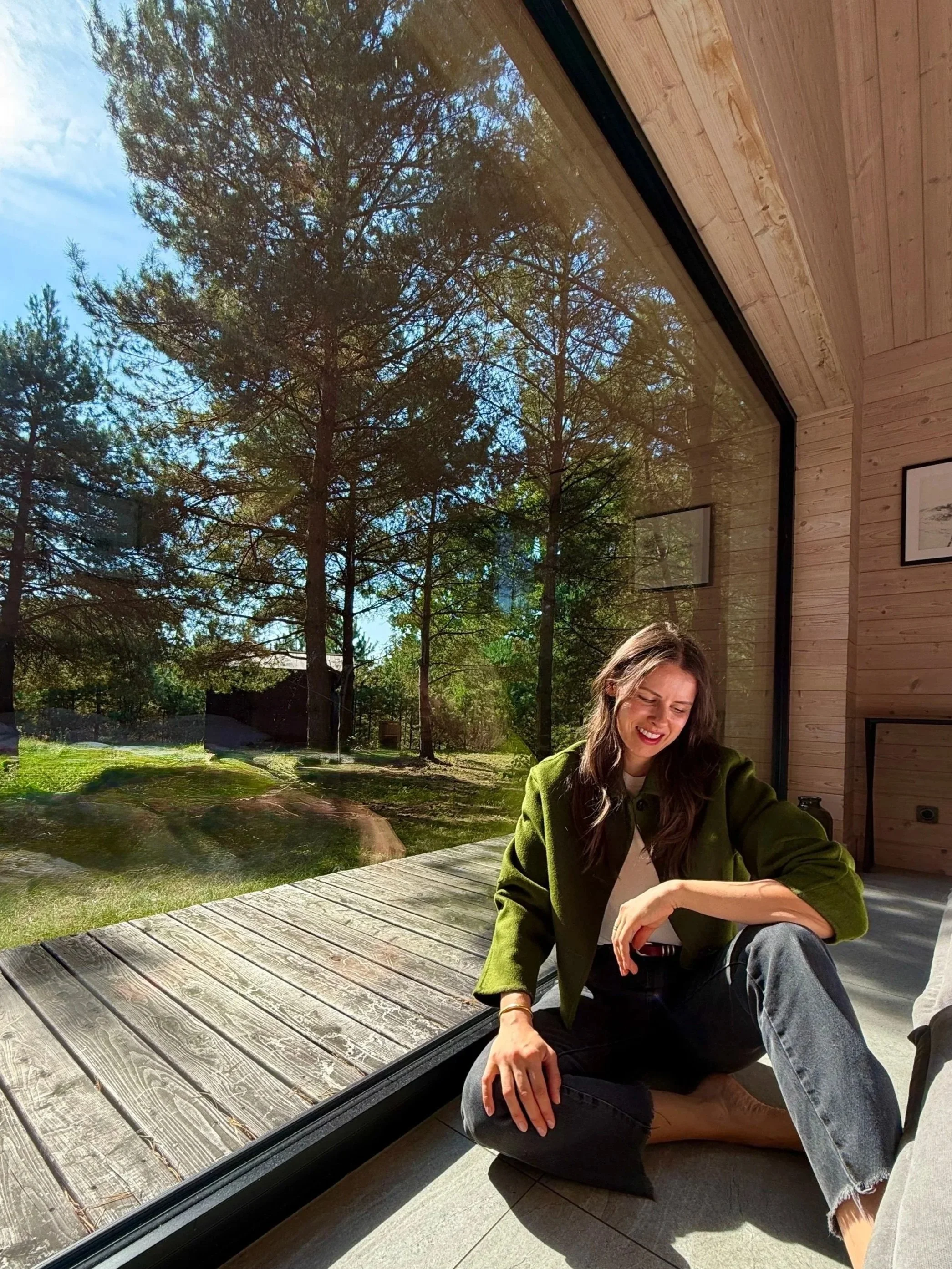 A woman with long brown hair sitting on a wooden floor next to a large window that looks out to a forested area with trees and blue sky. She is smiling, wearing a green jacket and dark jeans.