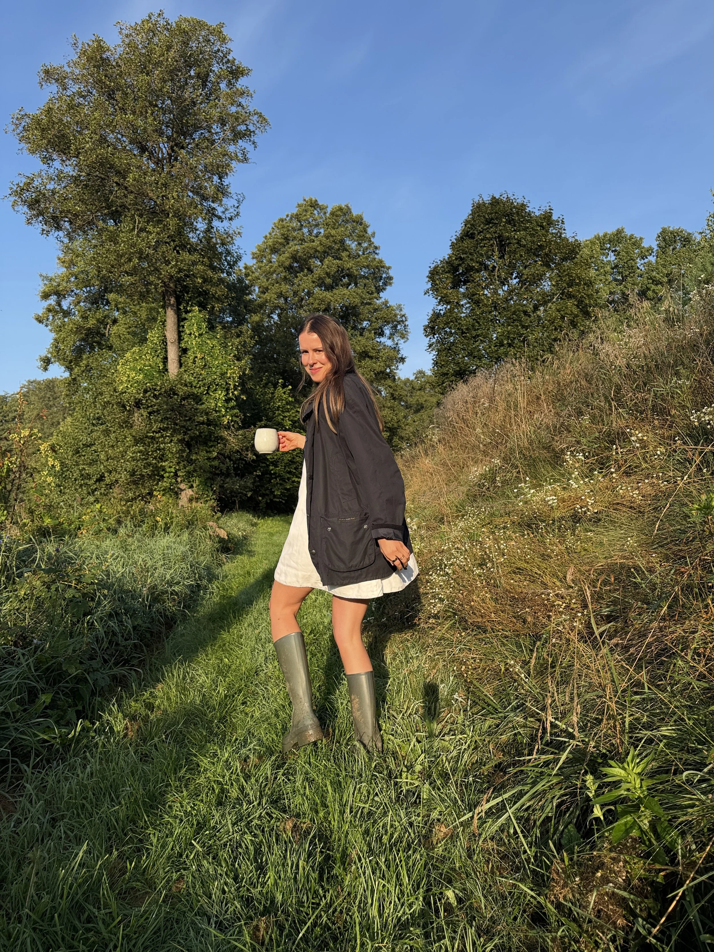 Anna Retel holding a mug and standing in a sunlit green meadow in a dark cardigan and rubber boots