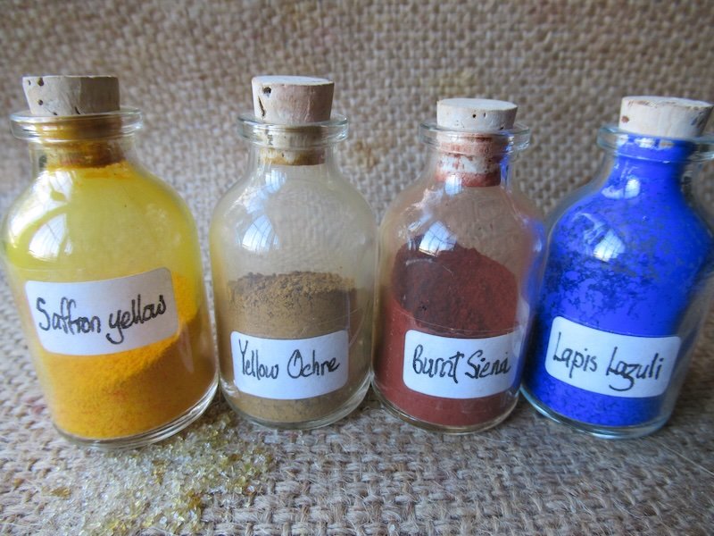 Four small glass bottles with cork stoppers containing different colored powders labeled saffron yellow, yellow ochre, burnt sienna, and lapis lazuli, placed on a textured surface.