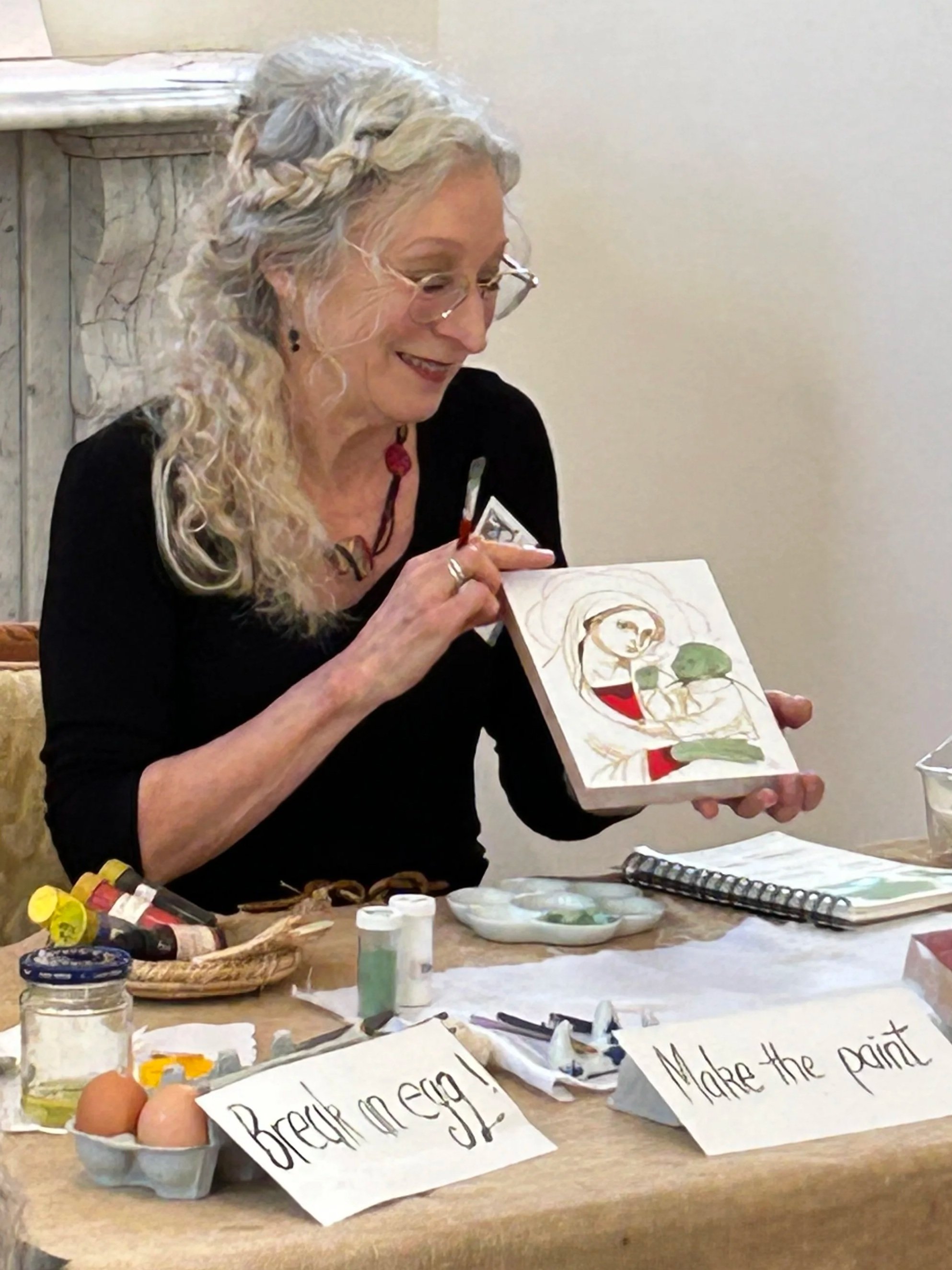 An elderly woman with long gray hair in a braid, wearing glasses, and a black shirt, is coloring a drawing of a child with a green head and red shorts. The table in front of her has art supplies and signs with handwritten text that says "Prepare your image!" and "More the paint".