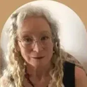 A woman with long, curly blonde hair wearing glasses, looking at the camera and smiling.