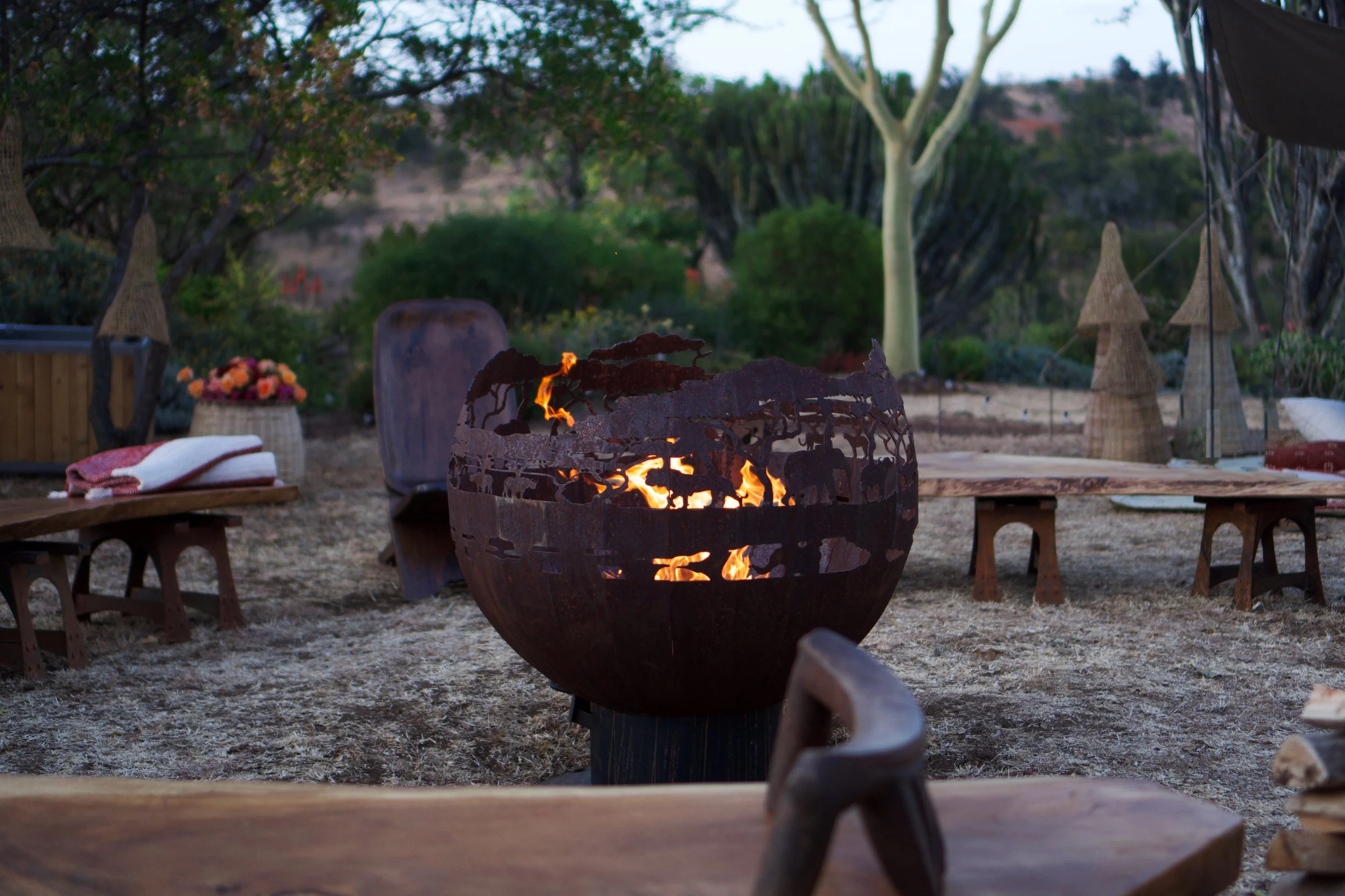Outdoor patio with a central firepit, surrounded by benches, towels, and decorations, with trees and a hillside in the background during dusk.