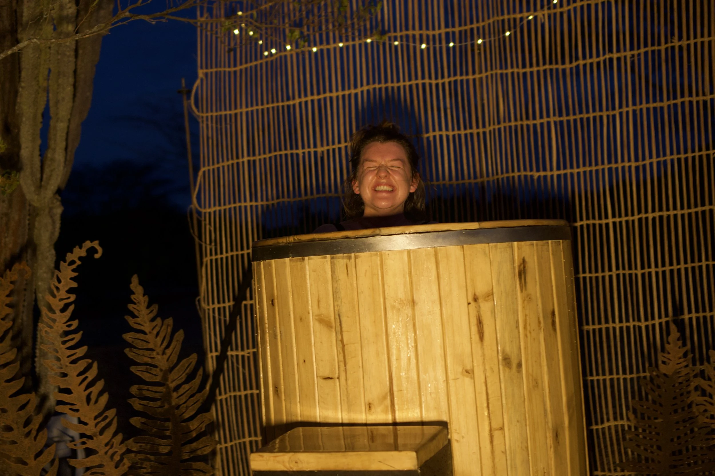 A person in a hot tub at night, surrounded by a bamboo privacy screen, with trees and string lights visible in the background.