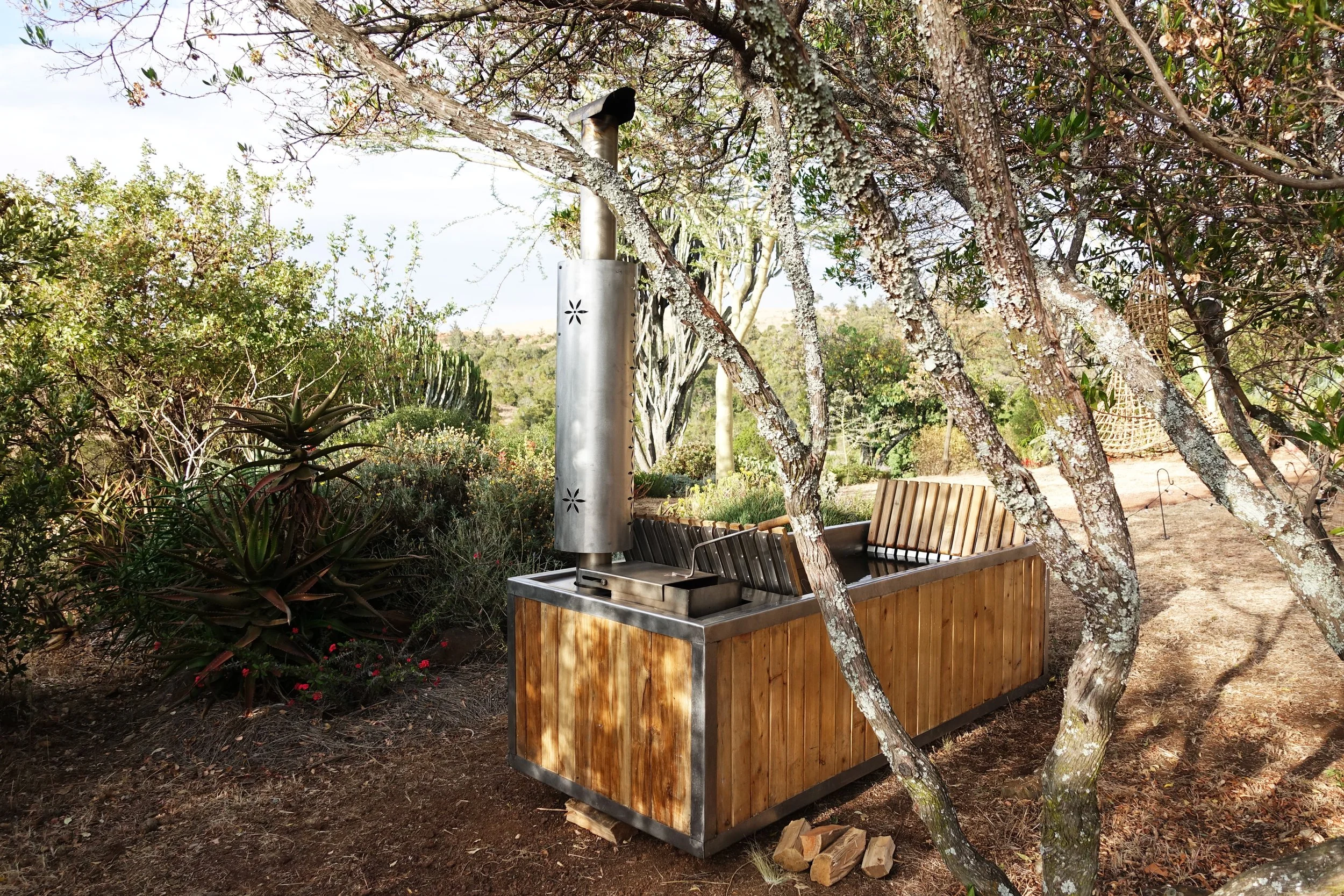 Outdoor scene with a wooden food truck, surrounded by trees and plants, with a metal chimney and a hanging wicker chair in the background.