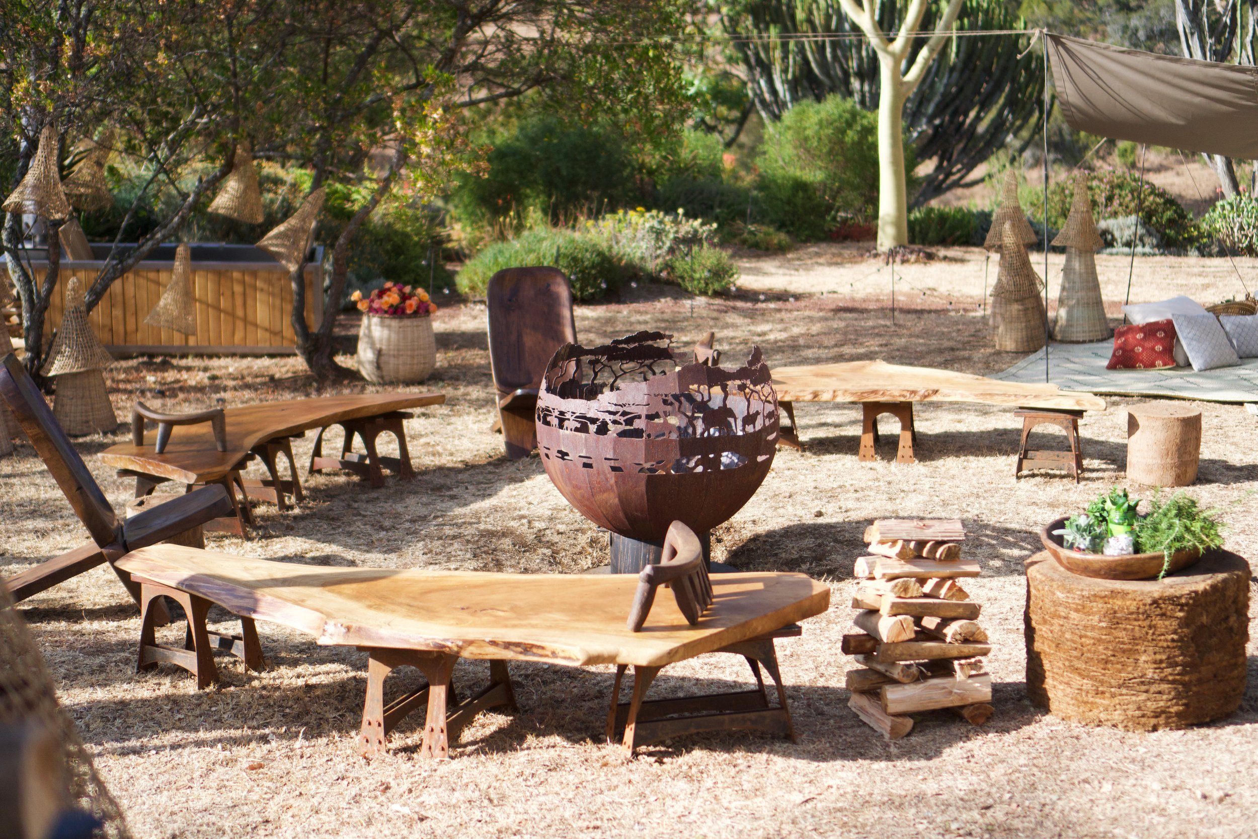 Outdoor garden scene with various wooden and wicker furniture, a fire bowl, potted plants, and trees, with a grassy and dirt ground.