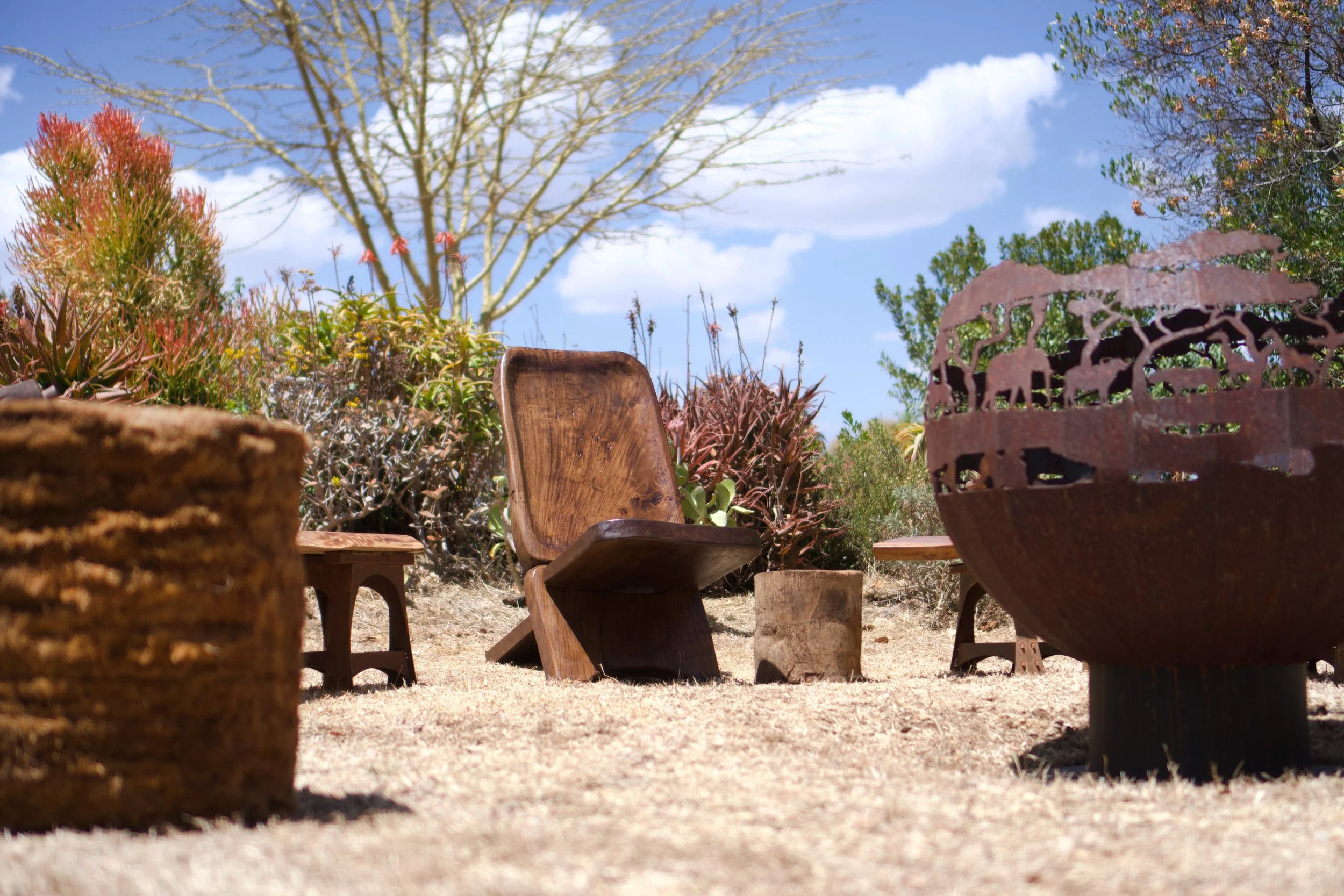 Outdoor seating area with wooden chairs and a metal fire pit, surrounded by desert plants and trees against a blue sky with clouds.