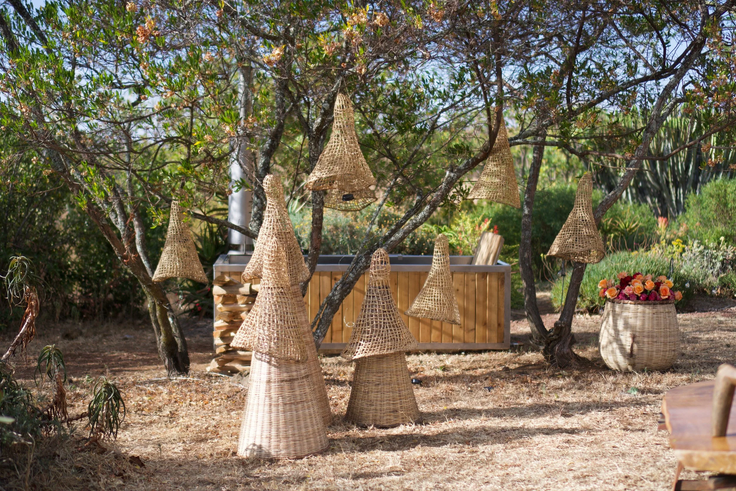 Wicker bell-shaped wind chimes hanging from tree branches in an outdoor garden setting, with a wooden bench and a basket of flowers in the background.