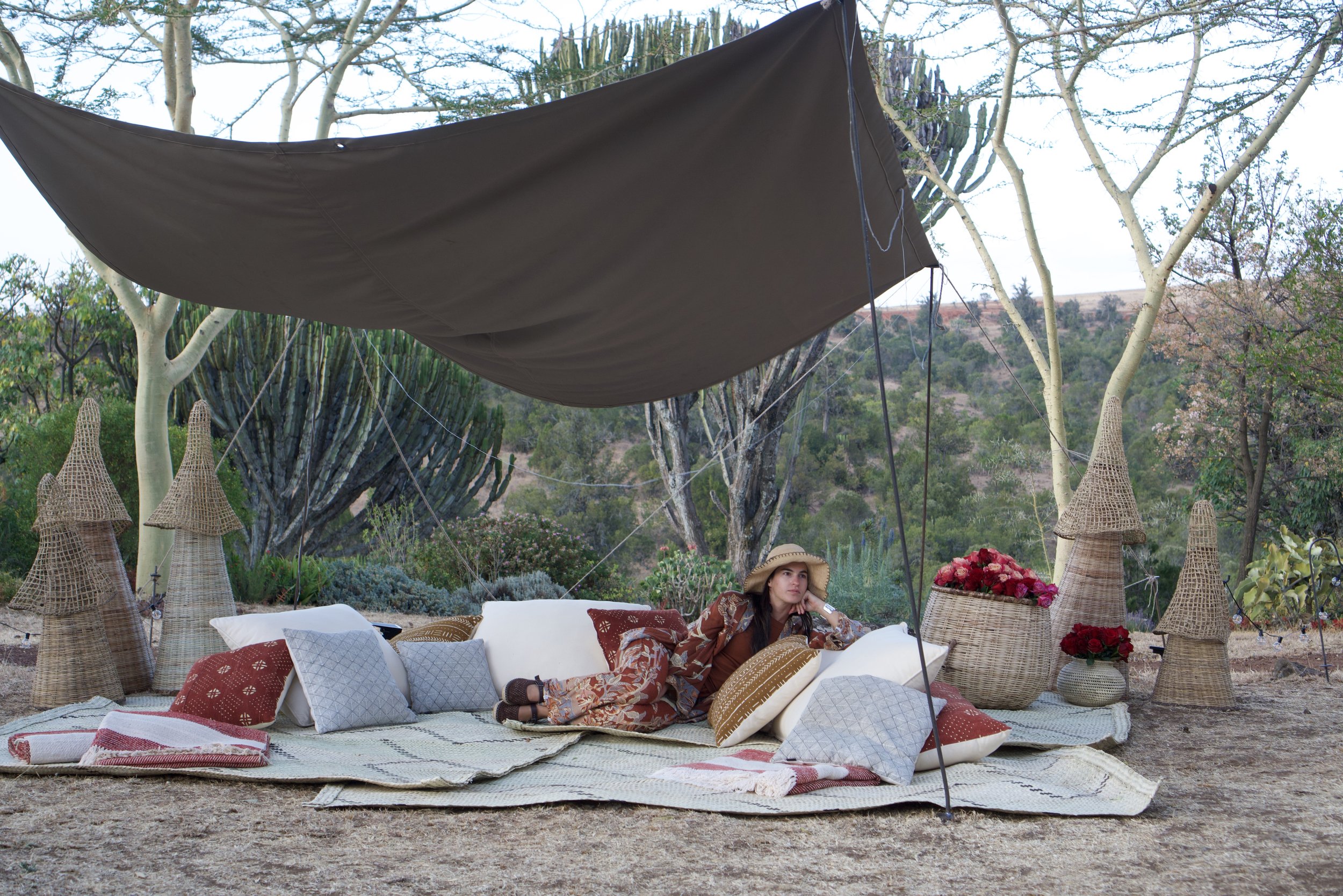 A woman dressed in bohemian style lies on a cozy outdoor seating area with cushions and throws, surrounded by wicker lamps and flowers, under a large shade sail amidst trees in a garden.