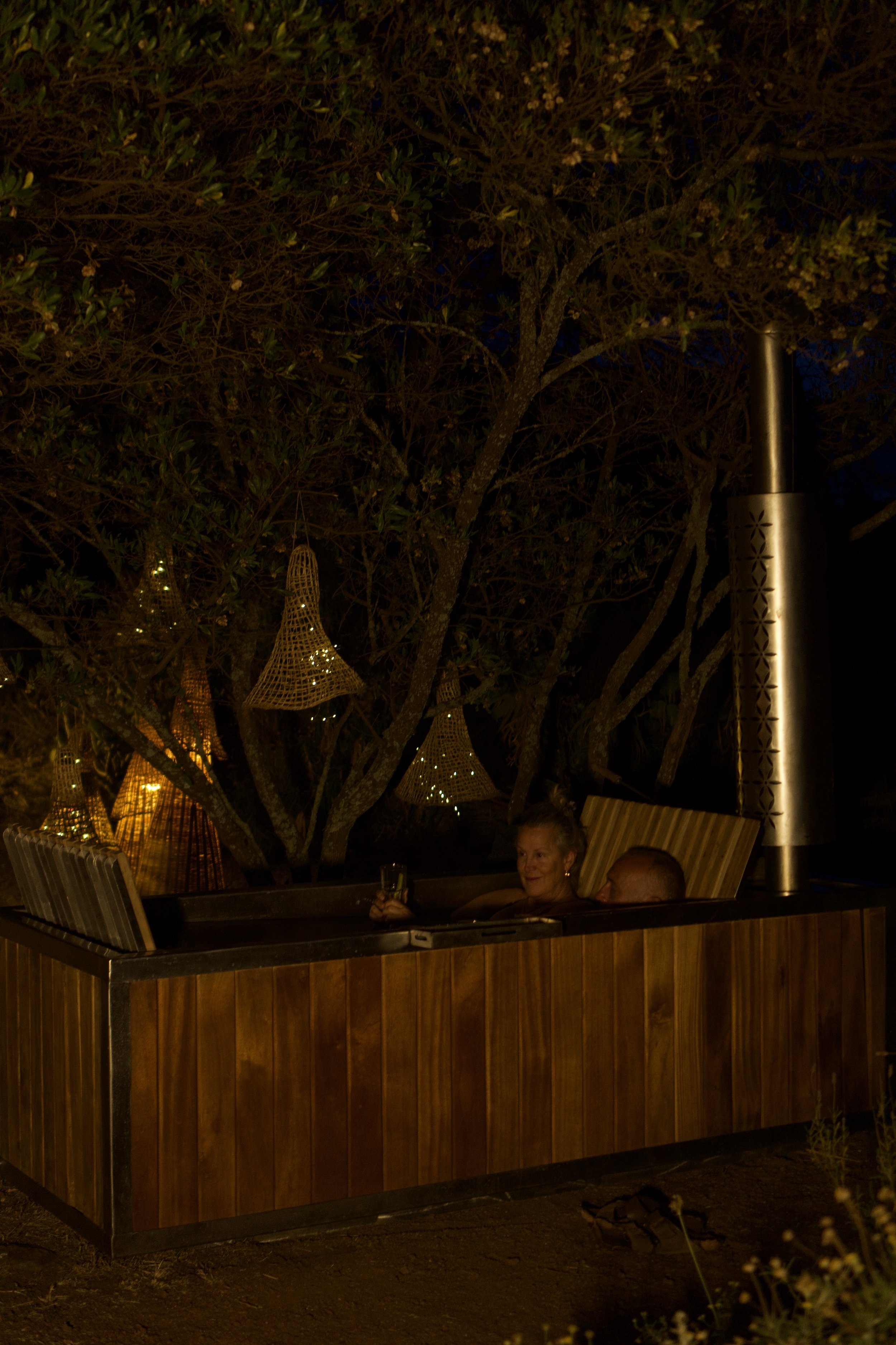 An outdoor hot tub at night with two elderly women inside, one holding a glass and smiling. The tub is surrounded by a wooden fence, and decorative hanging lanterns are in the background along with trees and a tall outdoor heater.