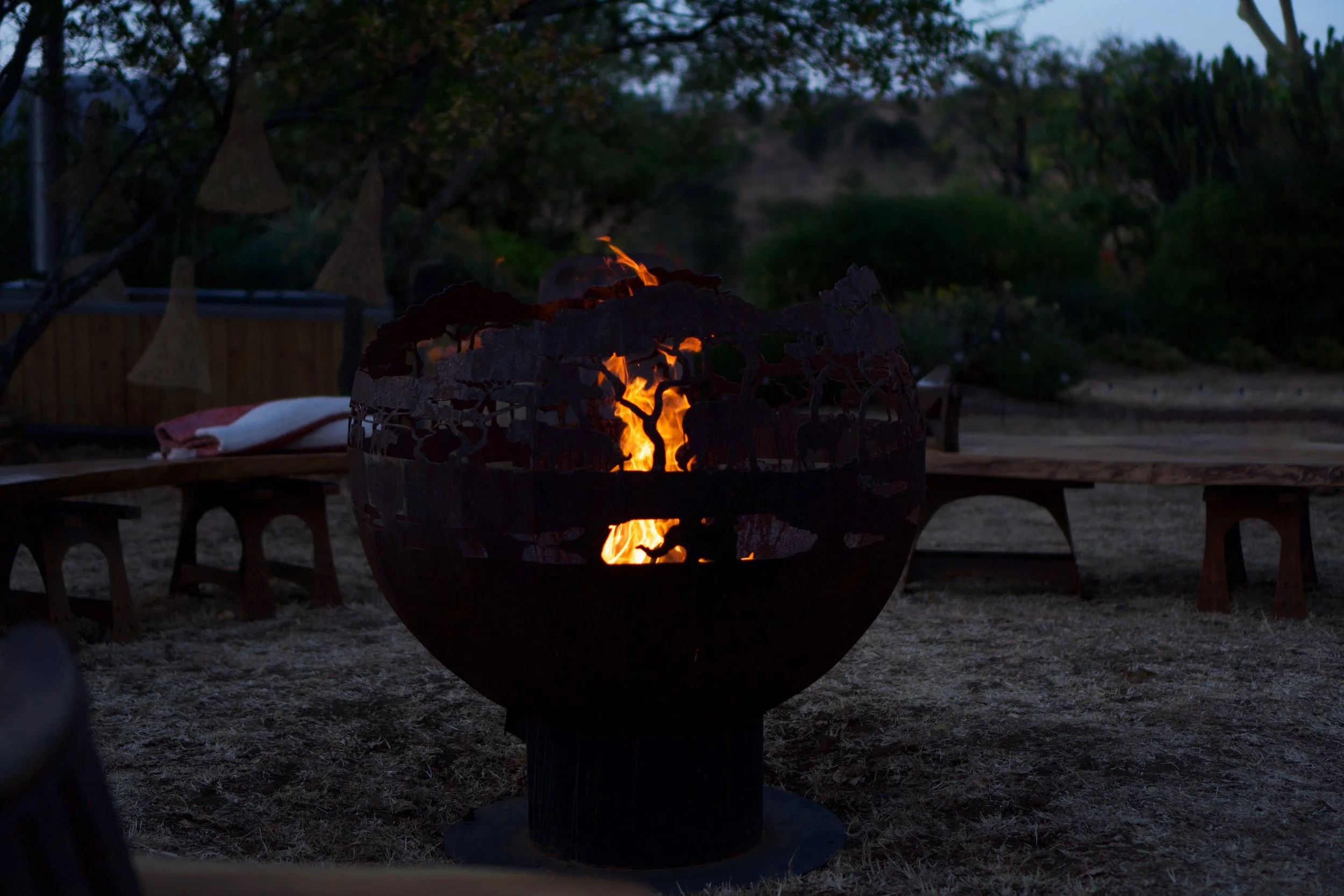 A fire burning in a metal fire pit in an outdoor setting during dusk or evening, with tables and trees in the background.