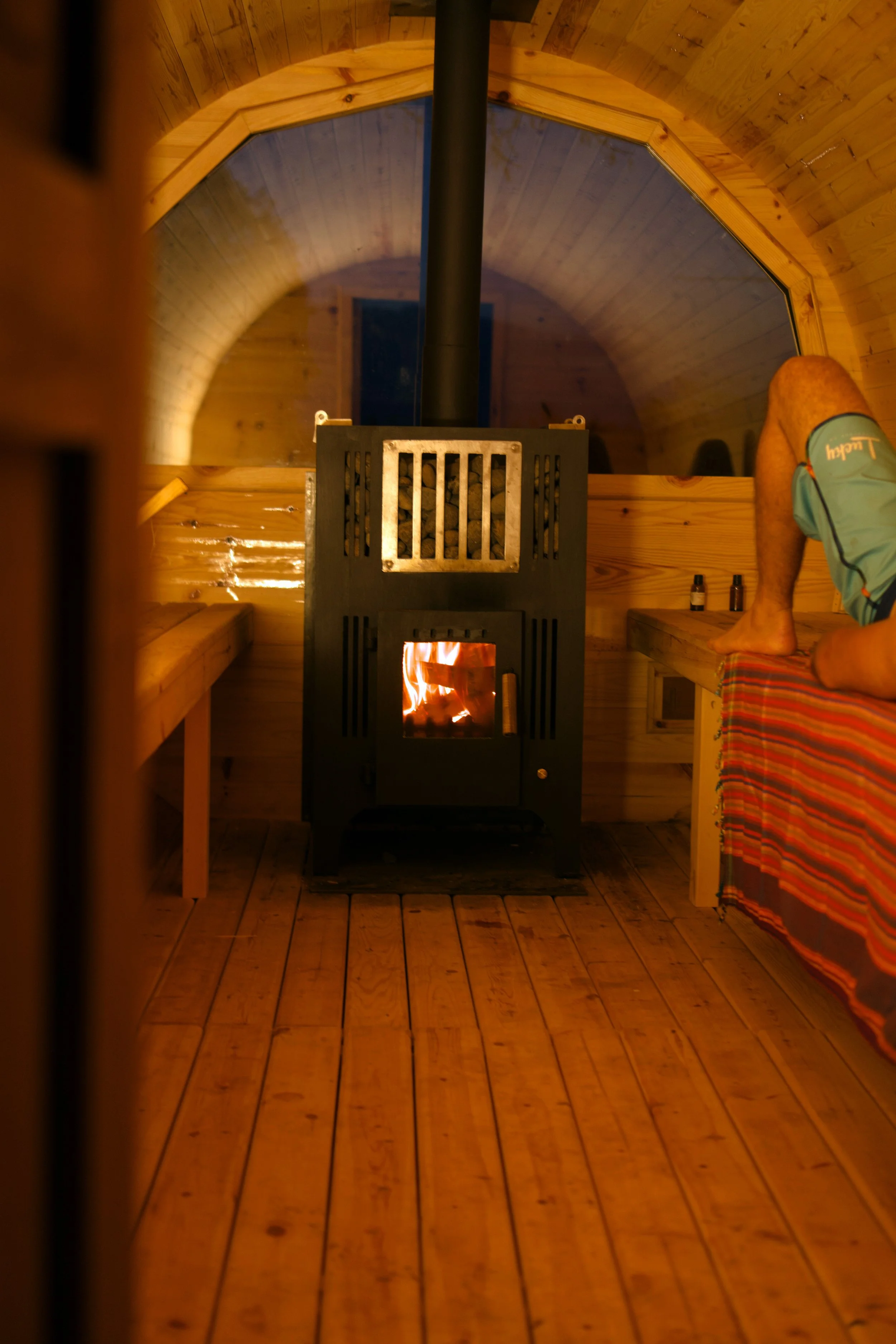 Interior of a small wooden sauna room with rounded ceiling, a wood stove with a fire burning, benches on either side, a person wearing plaid shorts and a blue shirt, and bottles on the bench.