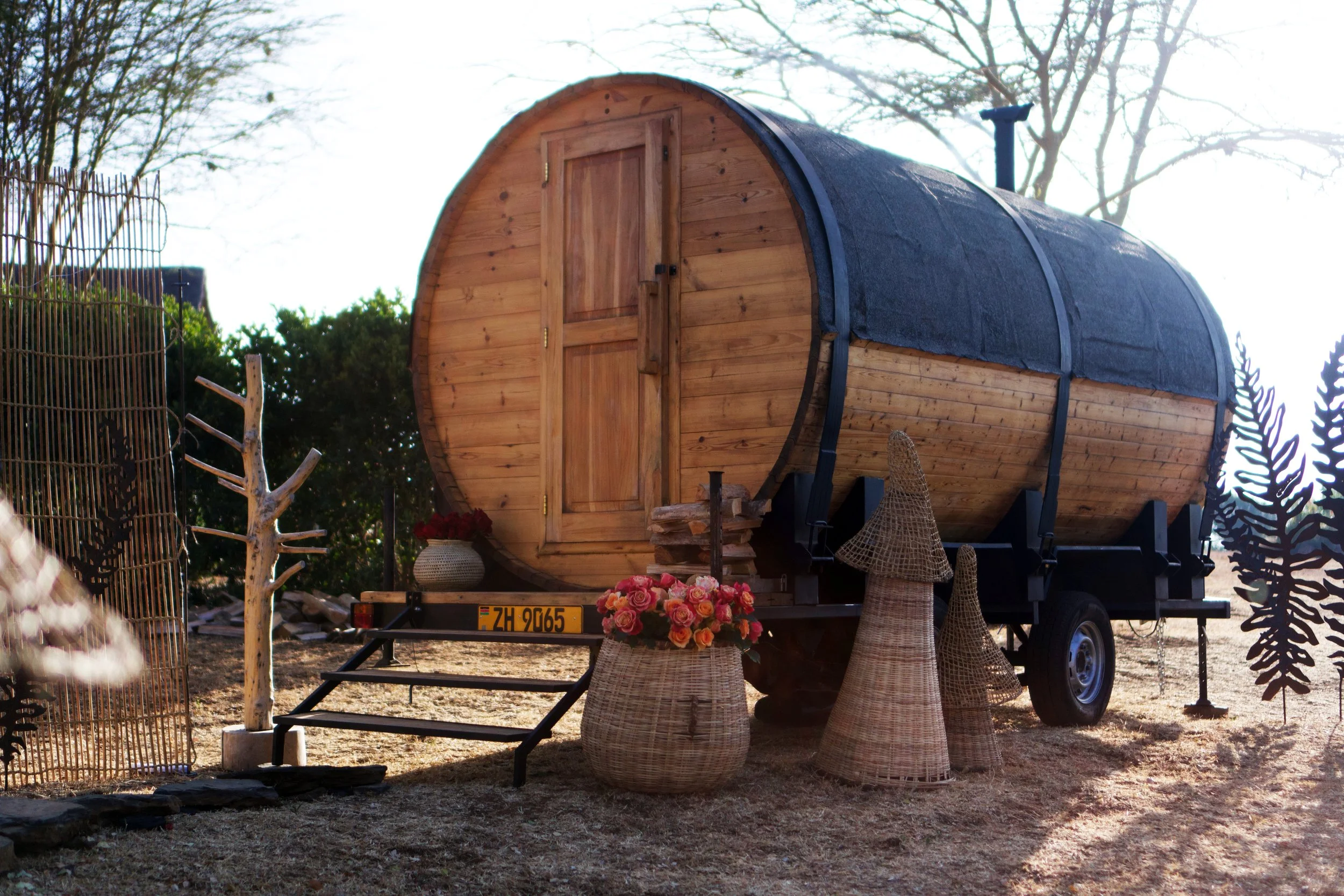A wooden tiny house on wheels with a rounded shape, situated outdoors with trees in the background. The house has a door on the front, a small set of stairs leading up to it, and decorative wicker baskets with flowers in front. There is a trailer hit