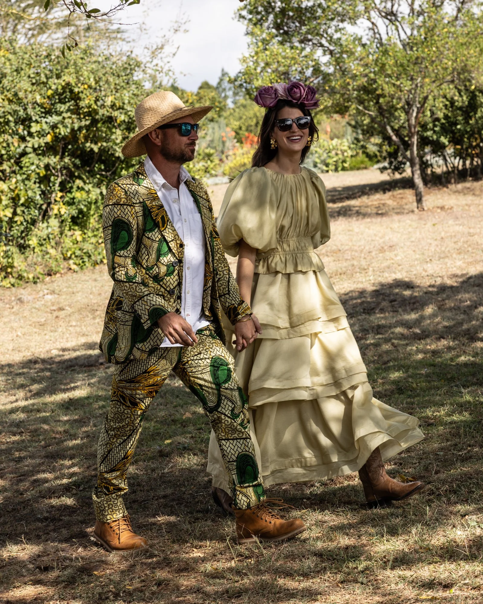 A man and woman walking outside in a park or garden, holding hands. The man is wearing a patterned blazer, matching pants, tan boots, a wide-brimmed hat, sunglasses, and a white shirt. The woman is wearing a cream-colored dress with puffed sleeves, large dark sunglasses, earrings, and a headpiece made of purple roses. The background features trees and green foliage.