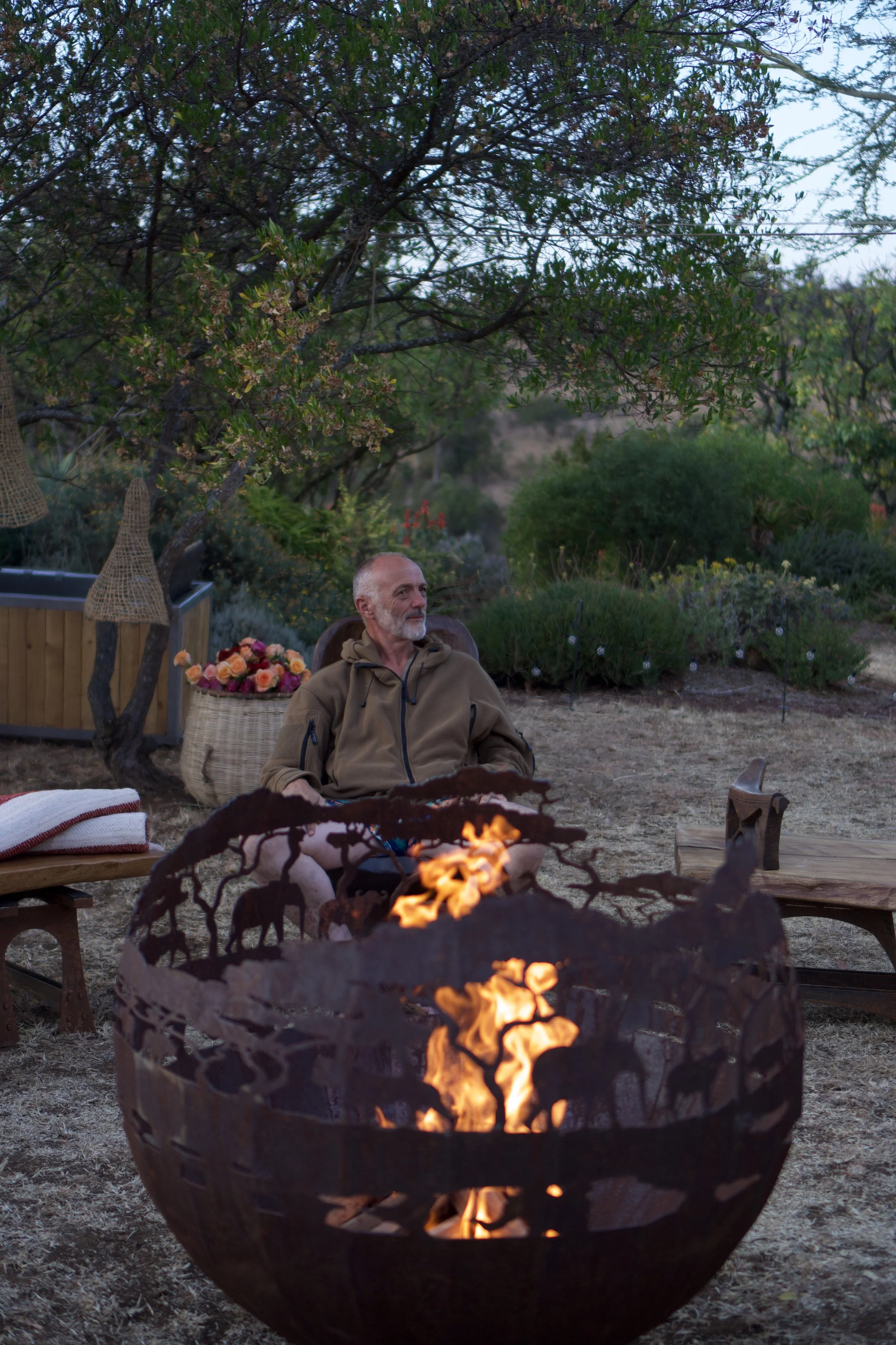 A man sitting outdoors near a fire pit with orange flames, surrounded by a garden with green bushes, trees, flowers, and outdoor decorations during dusk or early evening.