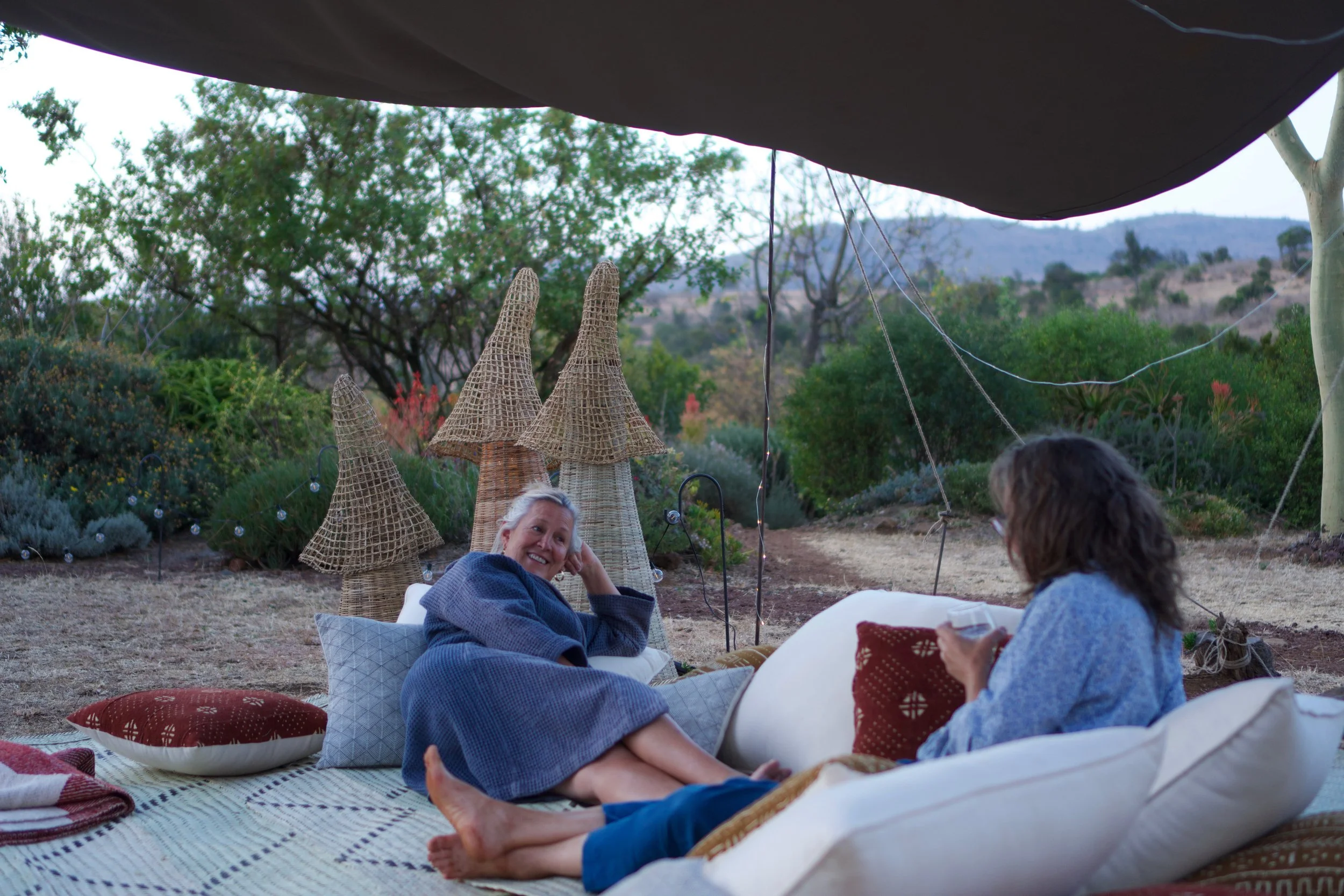 People relaxing outdoors under a large black canopy, sitting on cushions and blankets, with a scenic landscape of trees and hills in the background.